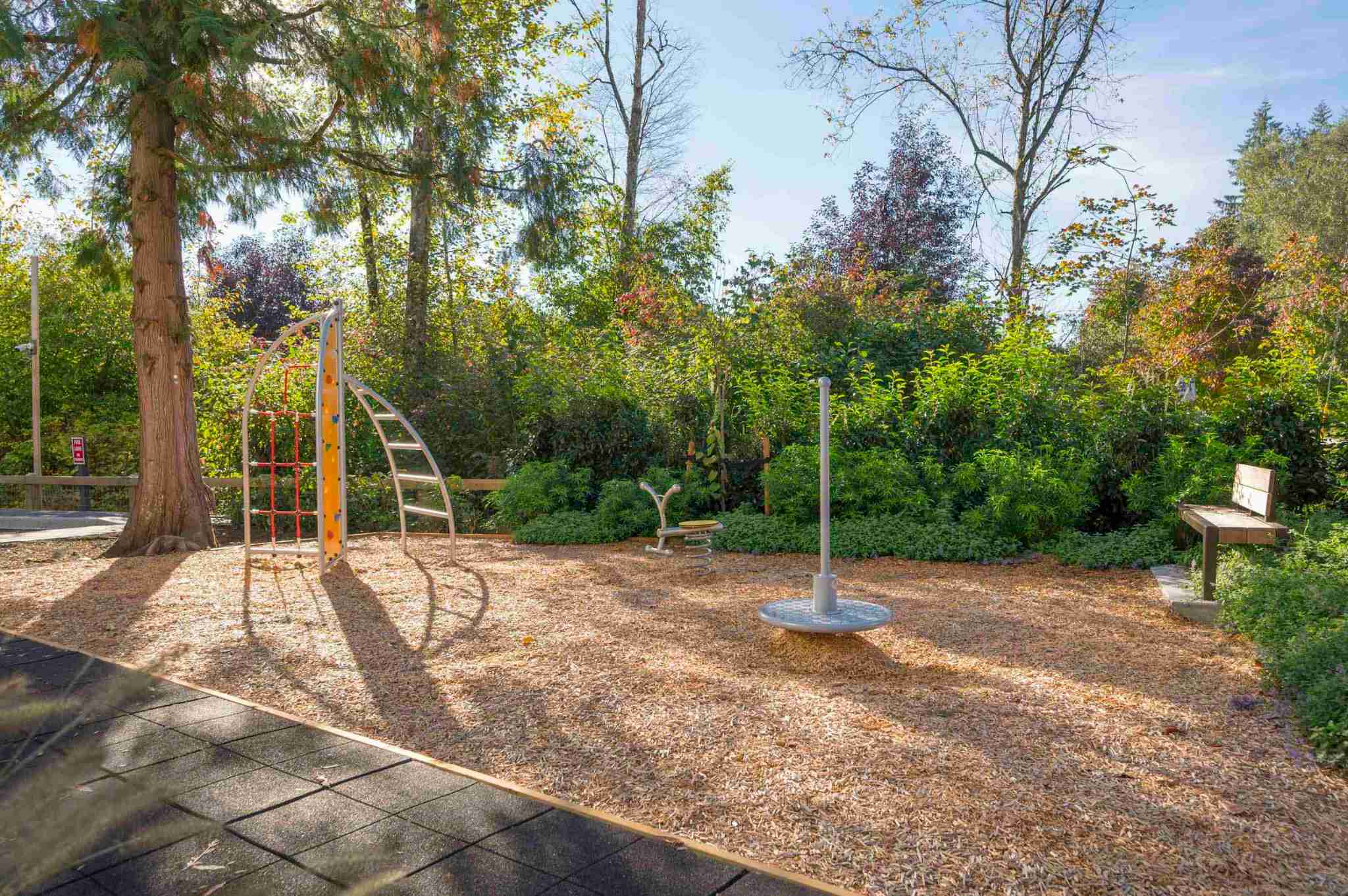 Park Avenue Townhomes – An outdoor children's play area with climbing structures, a spinner, bench seating, and surrounding trees.