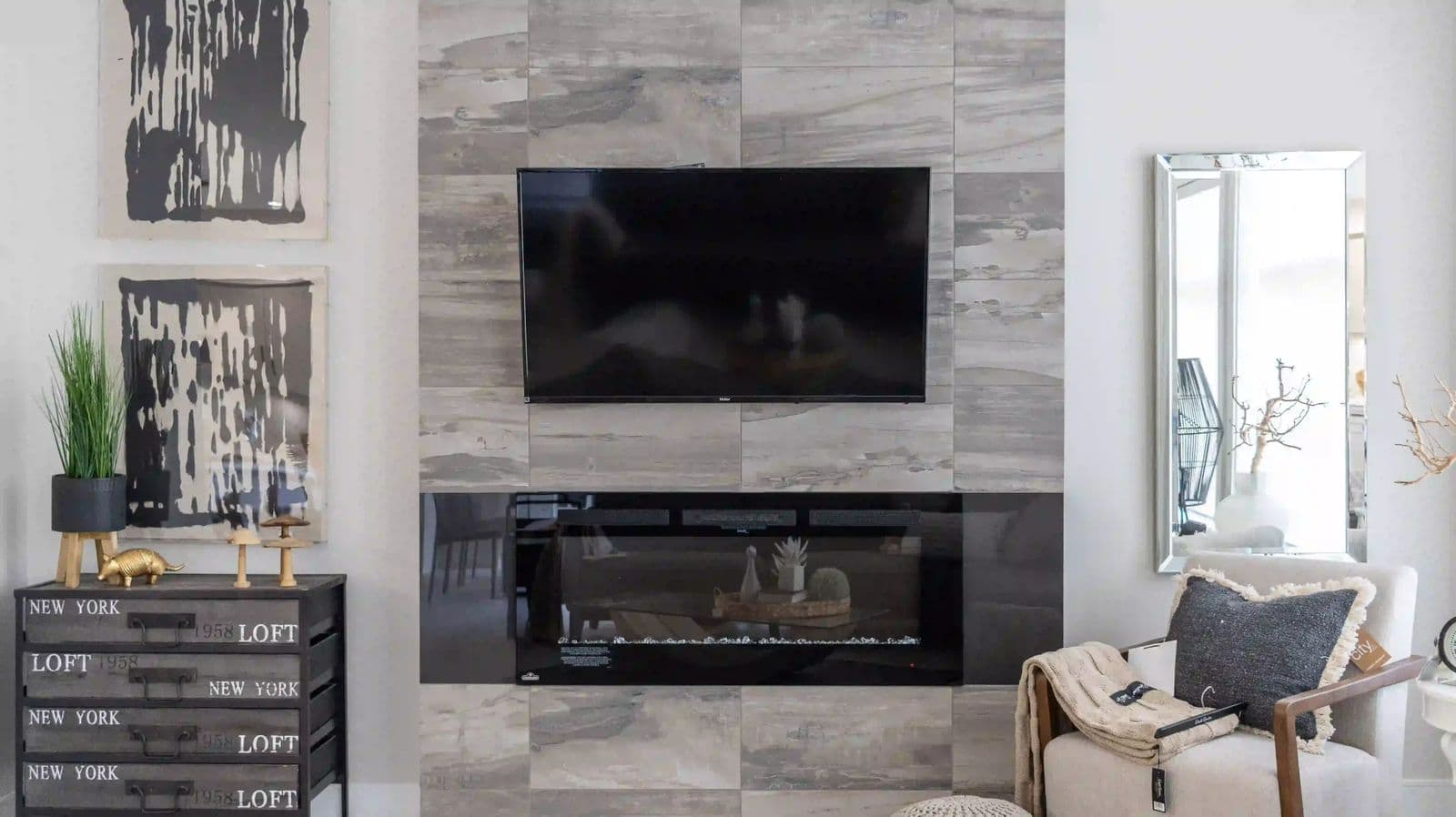 Liberty Encore – Living room with a wall-mounted TV above a built-in electric fireplace on a tiled accent wall, a dresser with decor, and a mirror to the side.