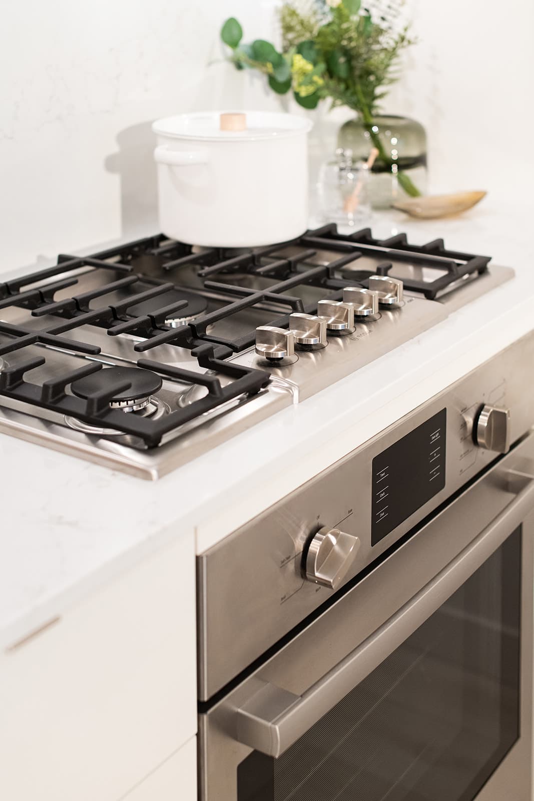 Jinju – A close-up view of a modern kitchen setup featuring a stainless steel gas cooktop with cast iron grates integrated into a white marble-patterned countertop, positioned above a matching built-in wall oven. A white ceramic cooking pot sits on the back burner, accompanied by subtle decorative styling including a green glass vase with foliage against the clean white backsplash.