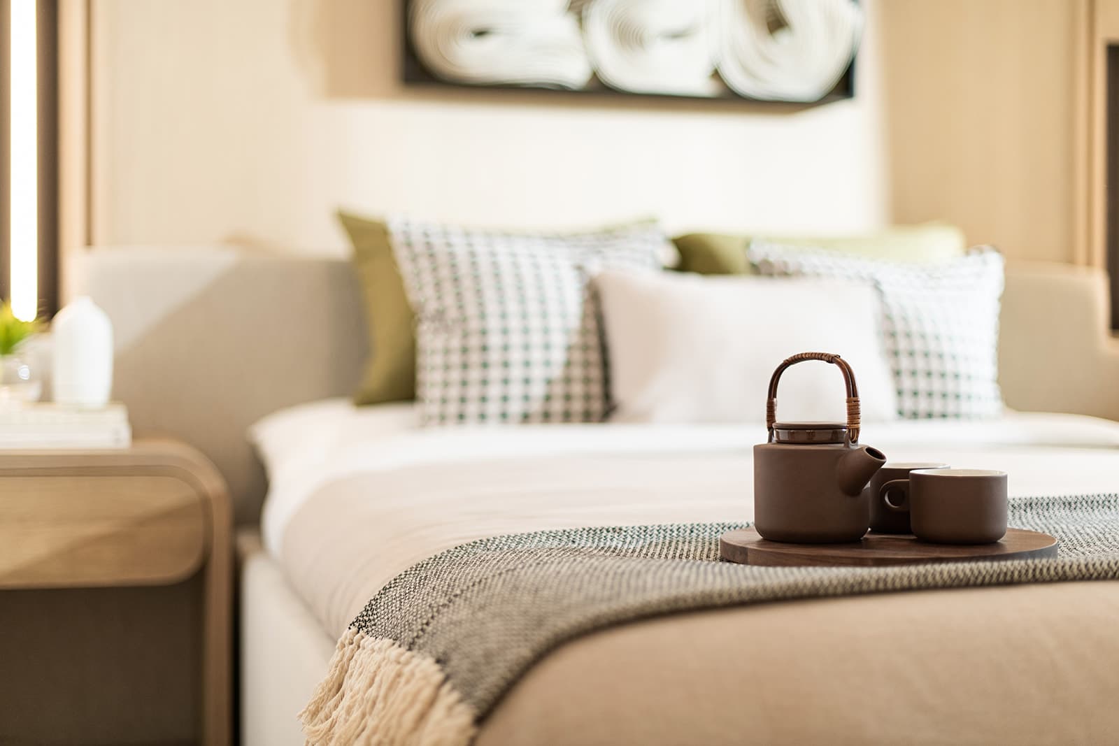 Jinju – A close-up view of a neatly made bed featuring a ceramic teapot and cups set on a wooden tray atop a textured throw blanket. In the soft-focus background, green and patterned accent pillows rest against an upholstered headboard alongside a light wood nightstand.