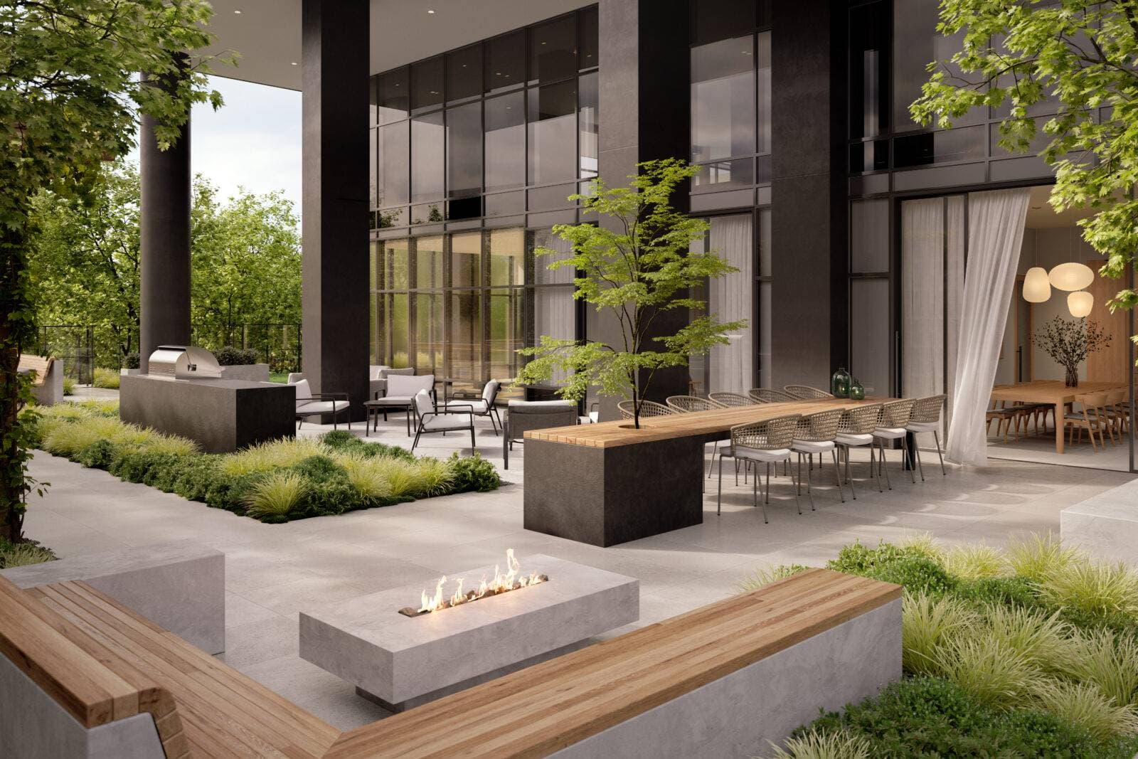 Jinju – A modern outdoor terrace amenity features a concrete linear fire pit surrounded by wood-topped benches, alongside a long alfresco dining table and an outdoor kitchen station. Set beneath tall dark pillars and floor-to-ceiling glass walls, the patio is framed by manicured garden beds and lush green trees in soft daylight.