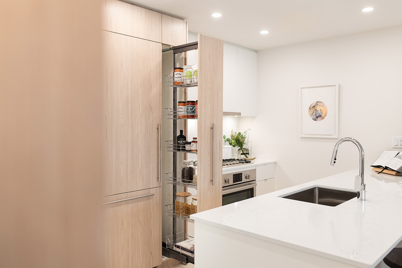 Jinju – A modern kitchen interior highlighting a tall, pull-out pantry cabinet with metal wire organizer shelves stocked with dry goods. The foreground features a white quartz island with an undermount sink and chrome faucet, set against light wood-grain and white cabinetry with stainless steel appliances.