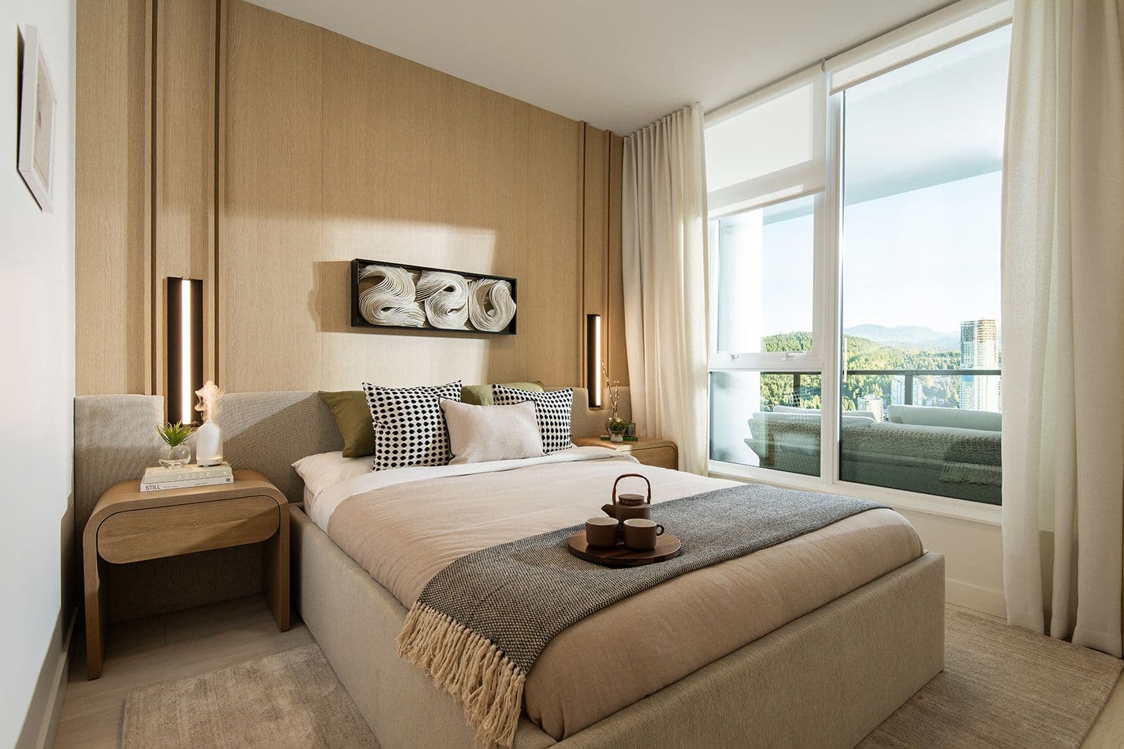 Jinju – A modern bedroom features a neatly made bed with light neutral linens and patterned throw pillows, set against a light wood-paneled accent wall with integrated vertical lighting. Floor-to-ceiling windows and sheer white drapes frame a bright daytime view of a balcony, distant forested hills, and high-rise buildings.