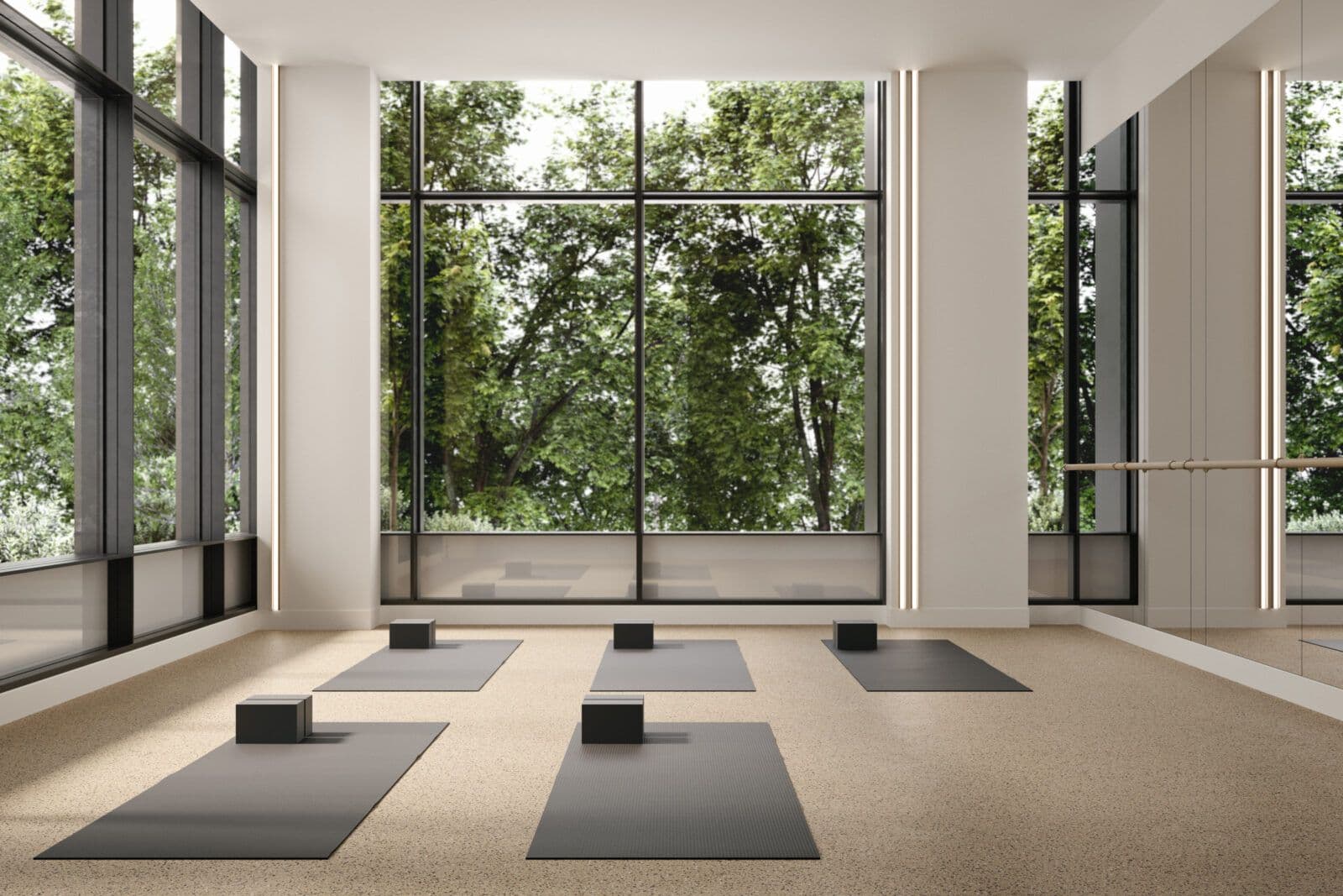 Jinju – A bright, minimalist yoga and fitness studio features speckled neutral flooring laid out with dark grey yoga mats and foam blocks. Floor-to-ceiling black-framed windows look out onto lush green trees, complemented by full-height wall mirrors, a ballet barre, and vertical light strips.