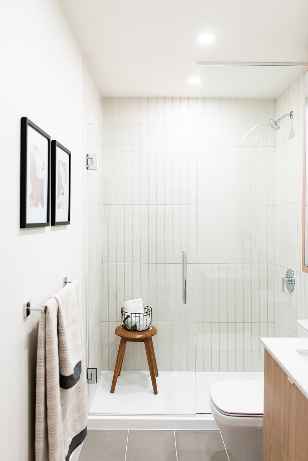 Jinju – A bright, modern bathroom featuring a walk-in glass shower lined with vertical white subway tiles, chrome hardware, and a small wooden stool inside. The surrounding space includes white walls decorated with framed art, a towel bar with a hanging towel, and a glimpse of a light wood vanity and toilet.