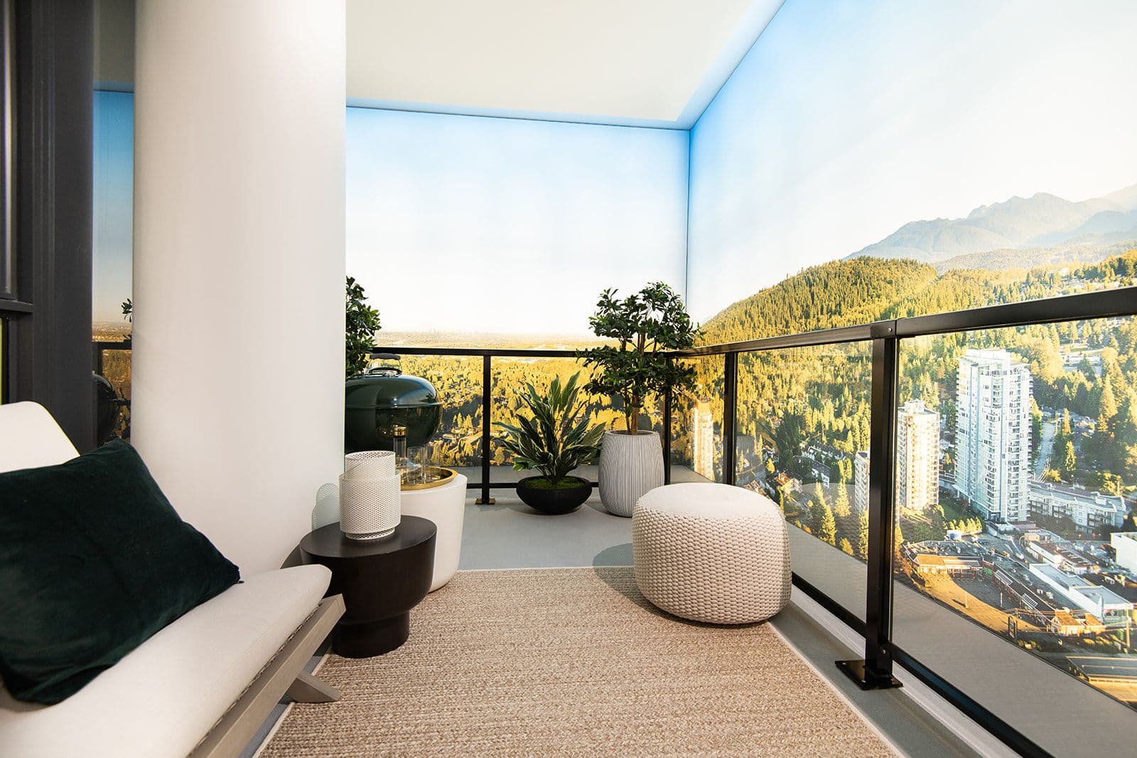 Jinju – A staged display suite balcony features a comfortable lounge chair, textured knit ottoman, small side tables, and potted plants on a neutral area rug. Glass railings overlook a realistic backdrop mural depicting a sunny, high-altitude view of forested mountains and distant residential towers.