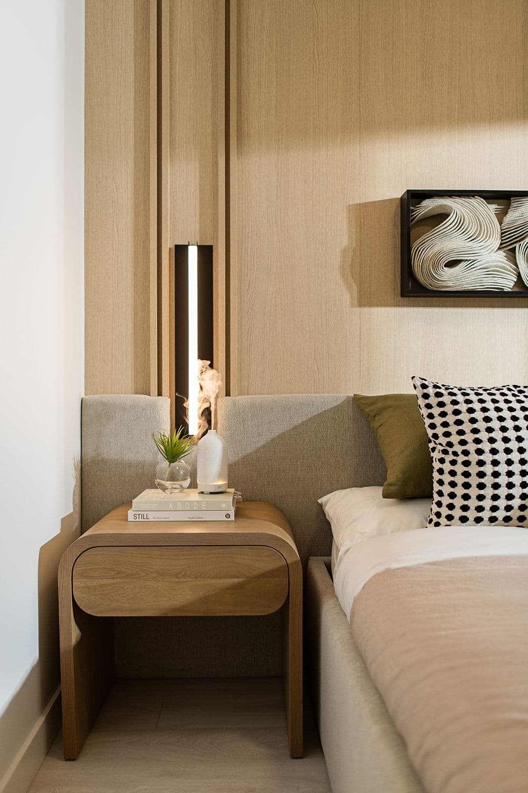 Jinju – A contemporary bedroom detail shot showcasing light wood wall paneling, an upholstered gray headboard, and a curved wooden nightstand with books and a small plant. A sleek vertical wall sconce and framed abstract dimensional art add warm, modern accents to the softly lit space.