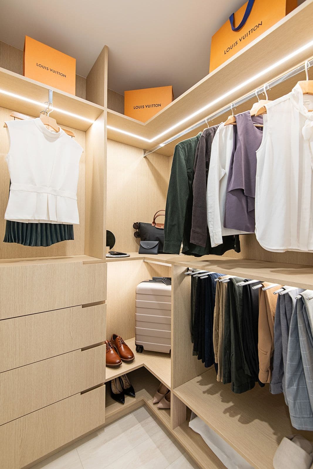 Jinju – A well-organized, modern walk-in closet made of light wood with integrated LED strip lighting along the shelves and hanging rods. It is neatly staged with hanging clothes, a row of folded pants, built-in dresser drawers, shoes, a suitcase, and designer shopping boxes on the upper shelves.