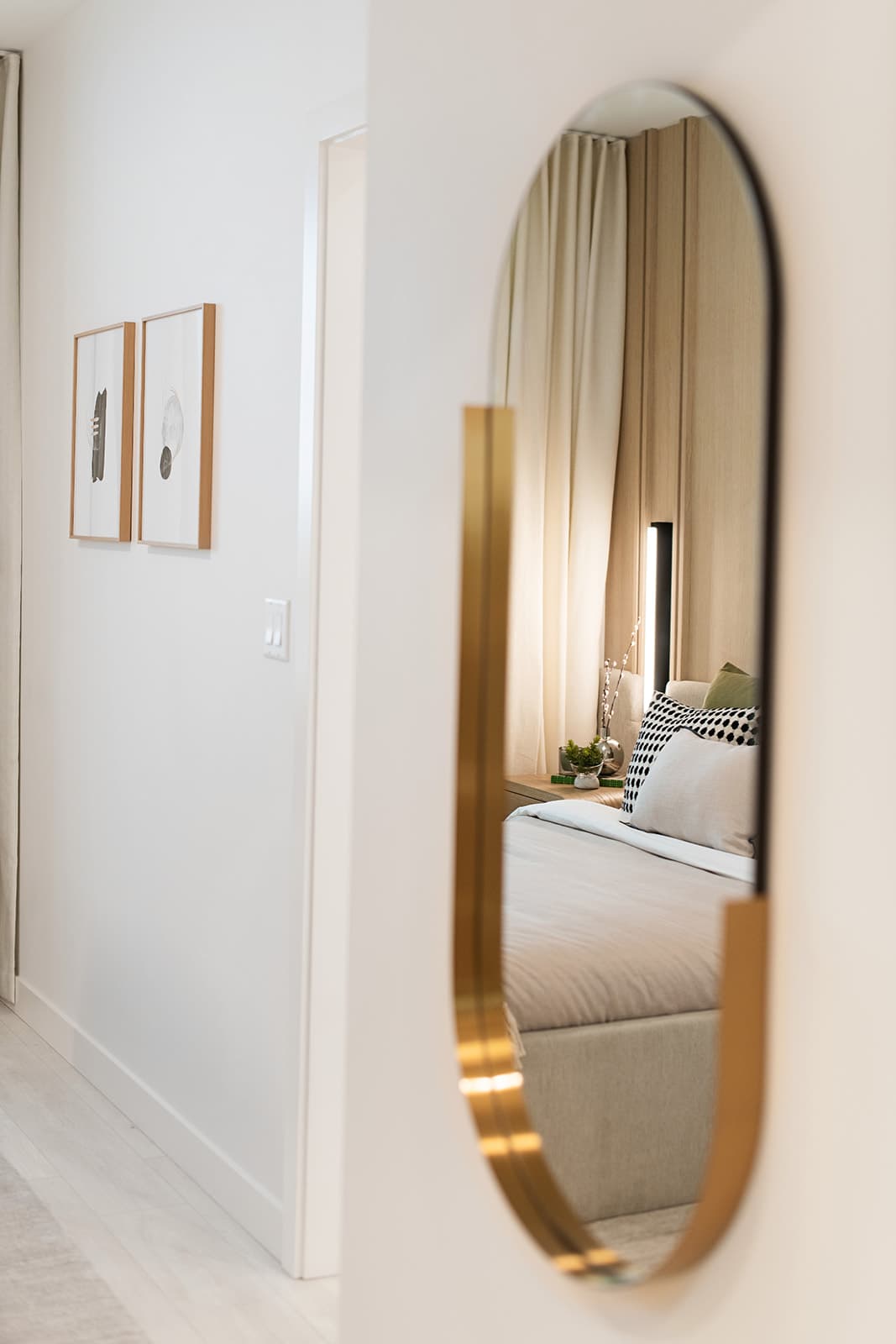 Jinju – A view down a bright white hallway features a brass-trimmed oval wall mirror that reflects a cozy, styled bedroom. Inside the reflection, a neatly made bed with neutral and patterned pillows sits beside a warm-lit lamp, sheer curtains, and wood-paneled accent walls.