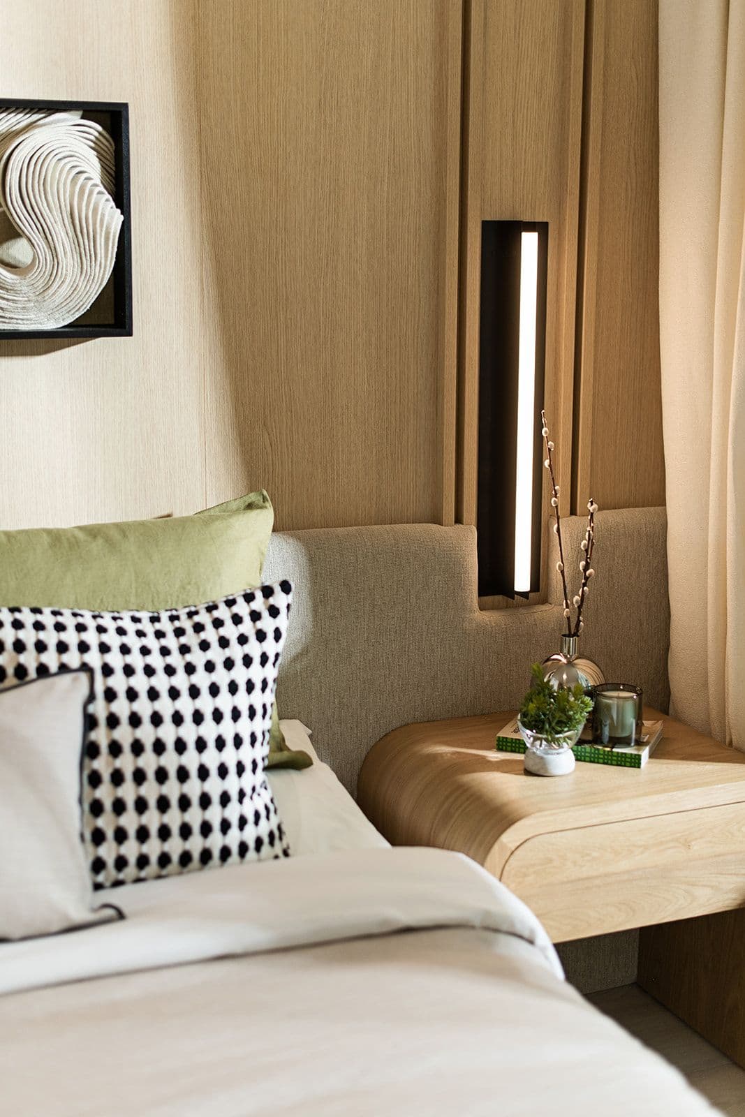 Jinju – A close-up view of a bedroom setup featuring a made bed with textured throw pillows, a custom fabric headboard, and wood-paneled walls. Next to the bed sits a curved light-wood nightstand with decor, illuminated by an integrated vertical wall sconce.
