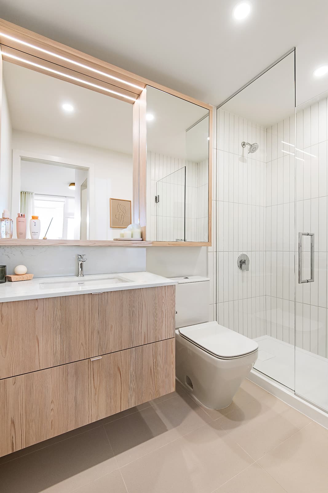 Jinju – A modern, bright bathroom featuring a light wood floating vanity with a white countertop and an integrated mirror medicine cabinet with warm under-cabinet lighting. A sleek toilet sits next to a walk-in glass shower lined with vertical white subway tiles.