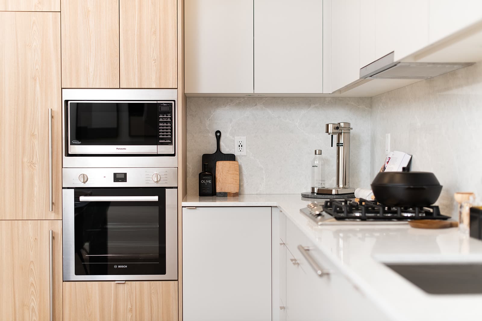 Jinju – A modern kitchen setup features built-in stainless steel appliances, including an integrated wall oven and microwave surrounded by light wood cabinetry. The adjoining white cabinets and light marble-patterned backsplash complement a clean countertop equipped with a gas cooktop, sleek sparkling water maker, and subtle cooking accessories.