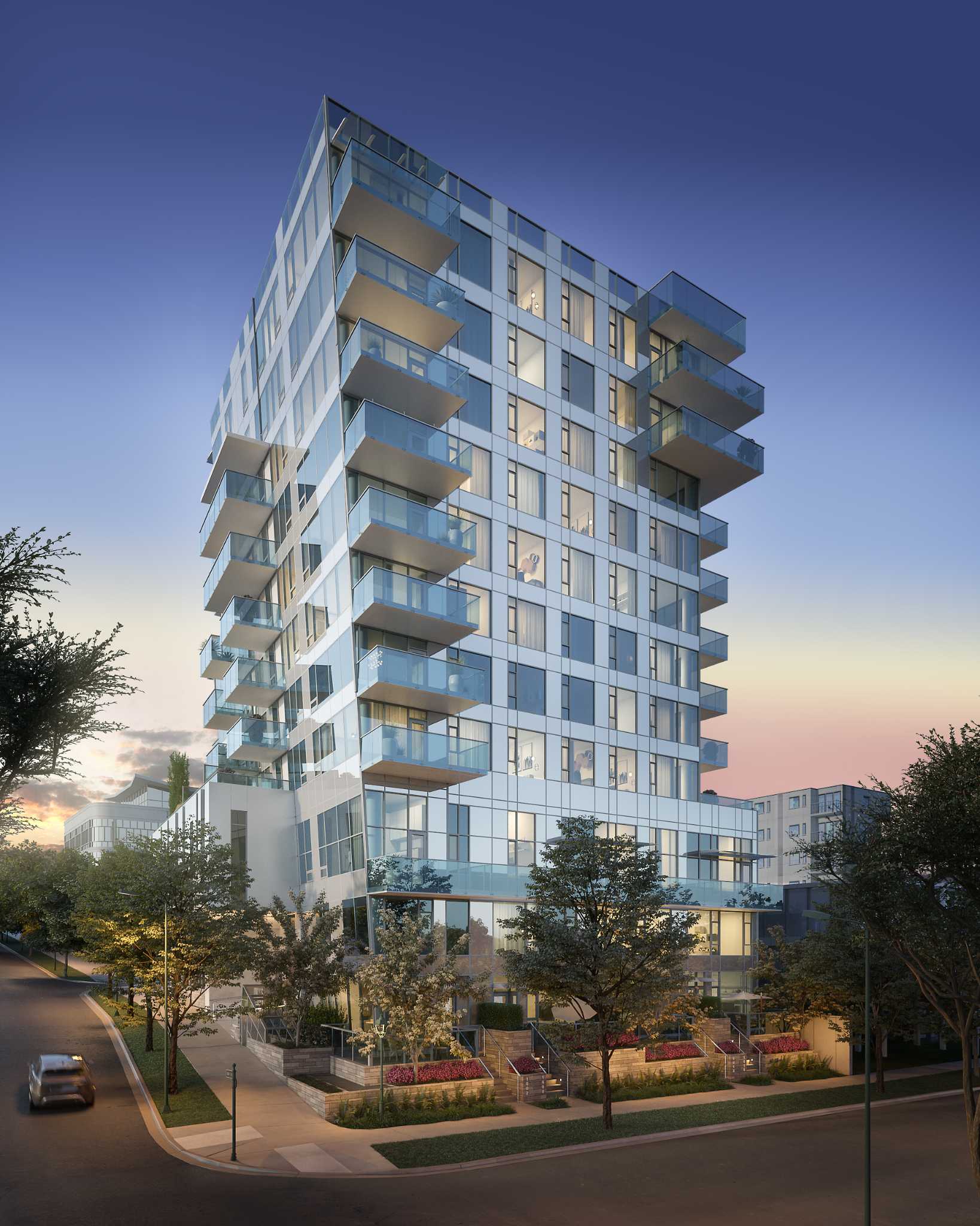 Italia – A glass-faced residential high-rise at dusk with rows of glass balconies, illuminated interiors, landscaped entrance planters, and surrounding street trees.
