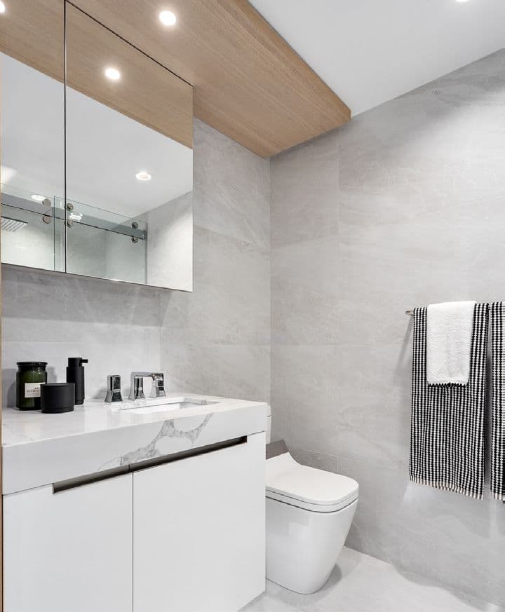 Italia – A bathroom with grey stone-look tile walls, a white marble vanity with black accessories, a wall-mounted toilet, and a striped towel hanging beside a mirrored medicine cabinet.