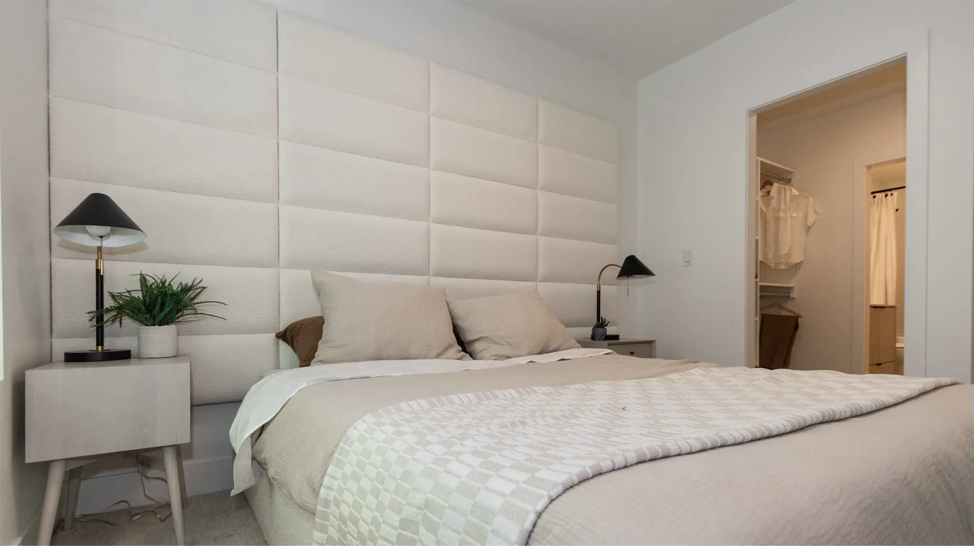 Elijah – A bedroom with an upholstered panel headboard wall, a made bed with neutral bedding, nightstands with lamps, and a walk-in closet and ensuite visible through an open doorway.