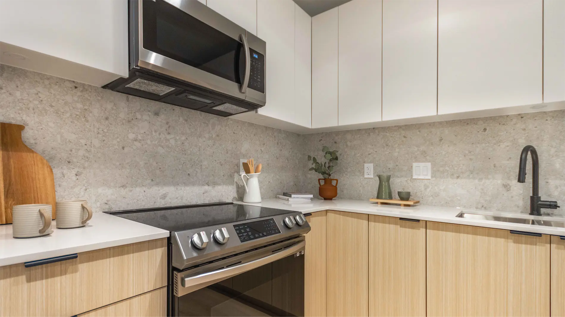 Elijah – A close view of a kitchen corner with white cabinetry, a stone-look backsplash, an over-range microwave, and mugs and a cutting board on the counter.