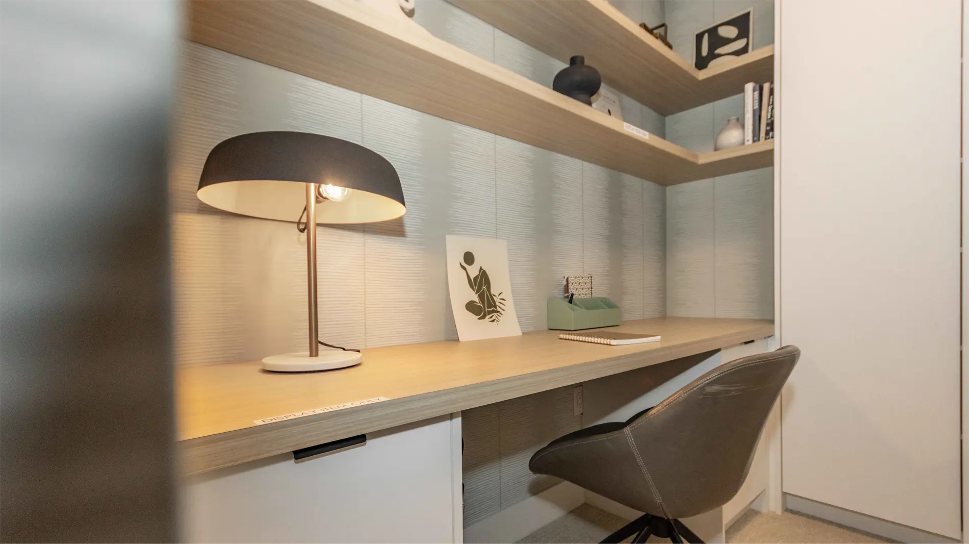 Elijah – A built-in desk nook with open shelving above, a black and brass desk lamp, decor items, and a grey office chair.