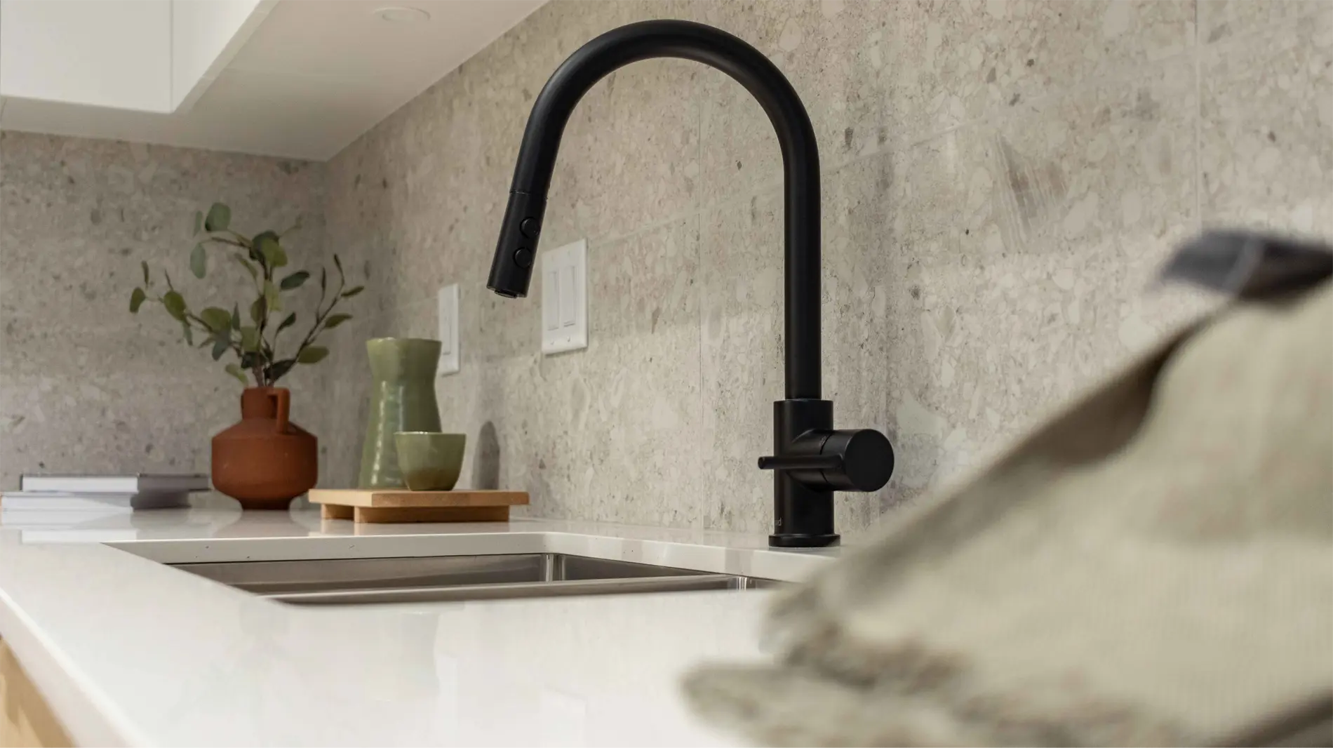Elijah – A close-up of a matte black kitchen faucet and undermount sink against a stone backsplash, with small decor vases on the counter.
