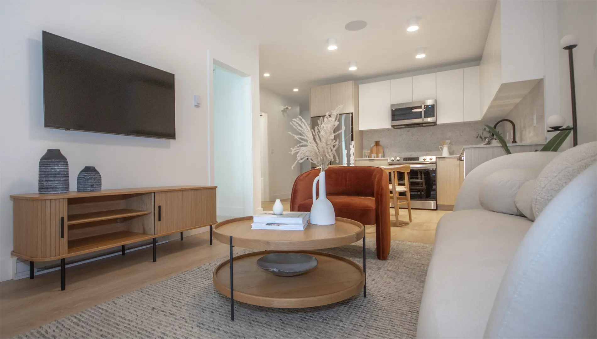 Elijah – An open-concept living room with a wall-mounted TV, wood media console, round coffee table, and a rust-coloured accent chair, with the kitchen and dining area visible beyond.