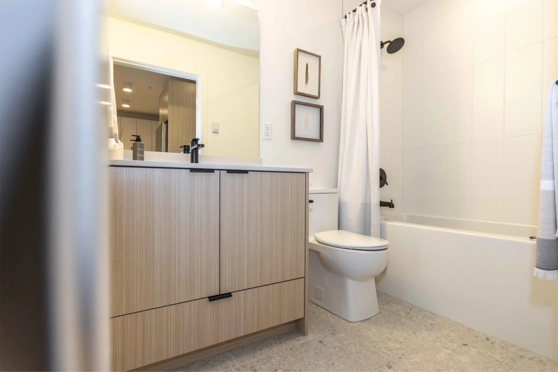 Elijah – A bathroom with a wood-look vanity, mirror, framed art, a toilet, and a tub-shower combo with a white curtain.