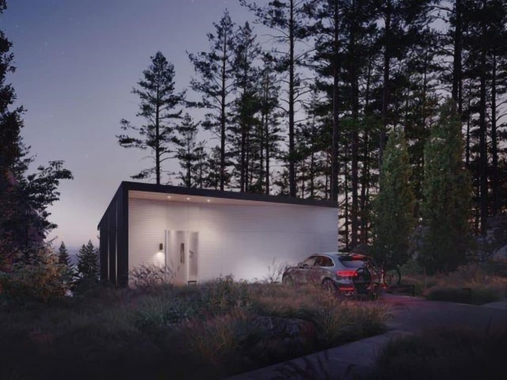 Alpine – Dusk exterior of a modern single-story home with dark and white siding nestled among tall forest trees, with a car parked in the driveway.