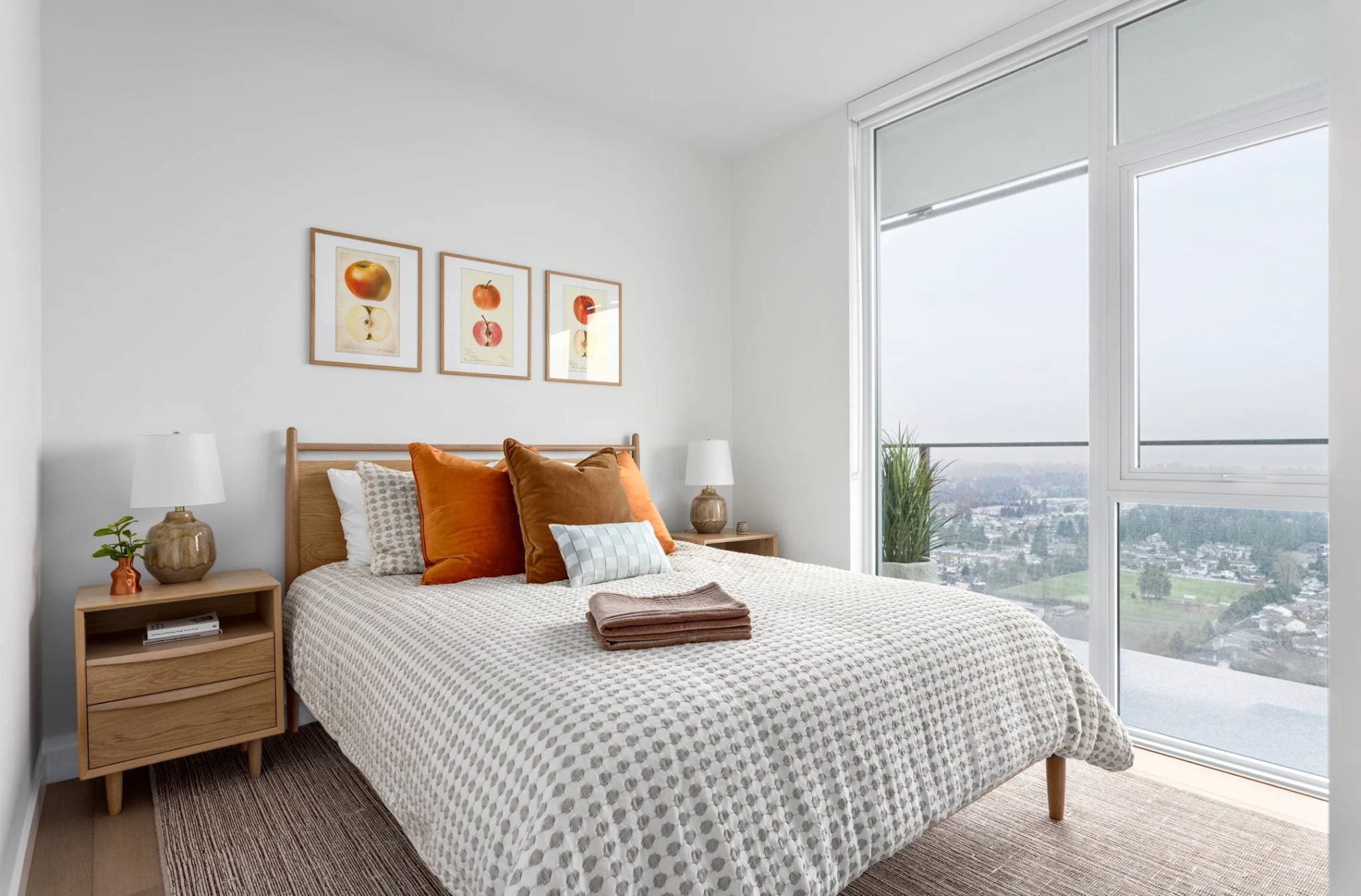 Century City Holland Park - Park Tower 1 – Bedroom with a wood headboard bed, orange and rust throw pillows, and a floor-to-ceiling window opening to a balcony overlooking a suburban landscape.