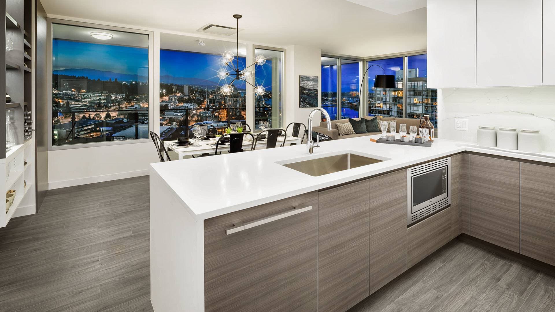 Ovation – An open-concept condo kitchen and dining space featuring a large island with white quartz countertops, wood-grain lower cabinets, and integrated stainless steel appliances. Floor-to-ceiling corner windows showcase a panoramic evening view of a lit city skyline and distant mountains.