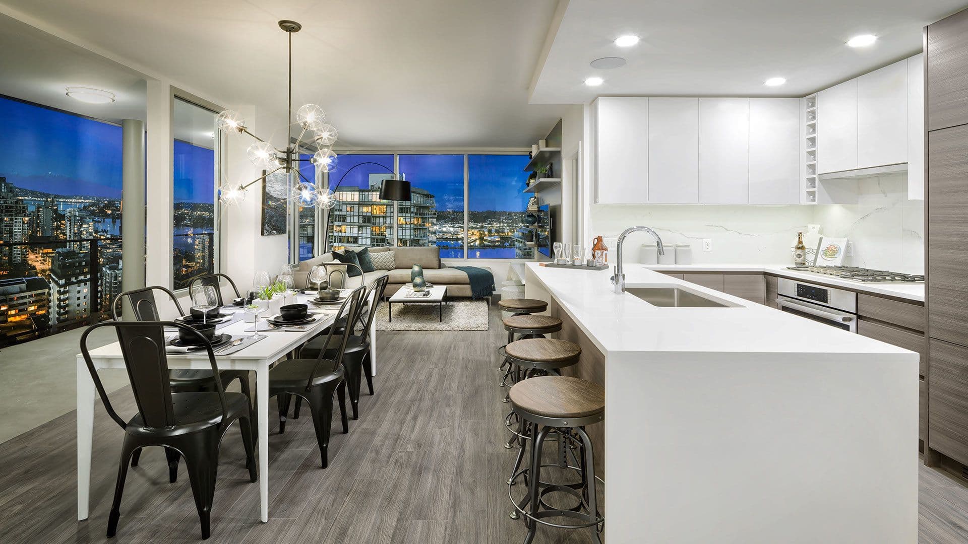 Ovation – An open-concept condo interior features a modern kitchen with a large white quartz island and a dining table set with black metal chairs under a multi-bulb sputnik chandelier. Floor-to-ceiling glass windows framed by dusk light reveal panoramic views of a illuminated city skyline and waterfront.