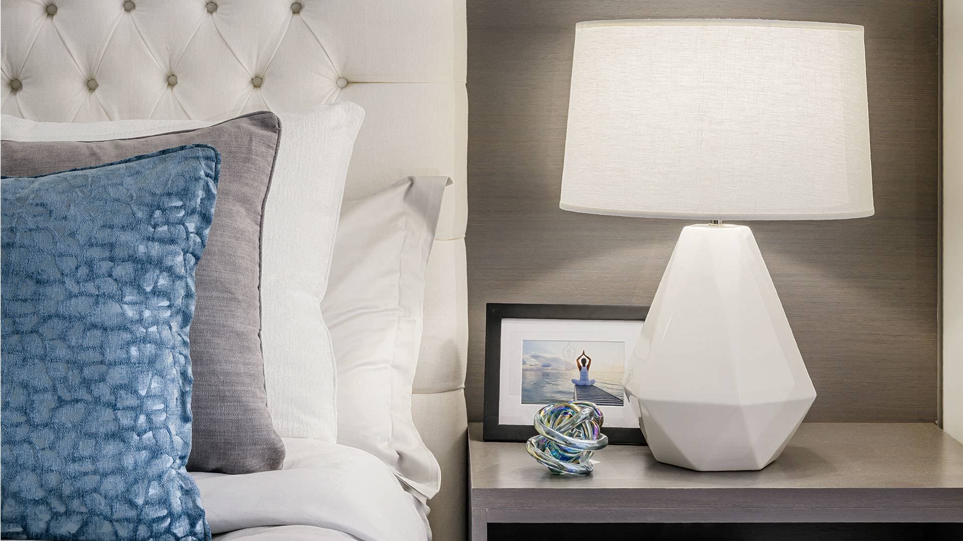 Ovation – A close-up shot of a modern bedroom nightstand featuring an illuminated geometric white table lamp alongside an upholstered tufted headboard and layered pillows in shades of blue, gray, and white. A small framed picture and decorative glass sculpture sit on the dark wood nightstand surface, creating a cozy and elegant mood.