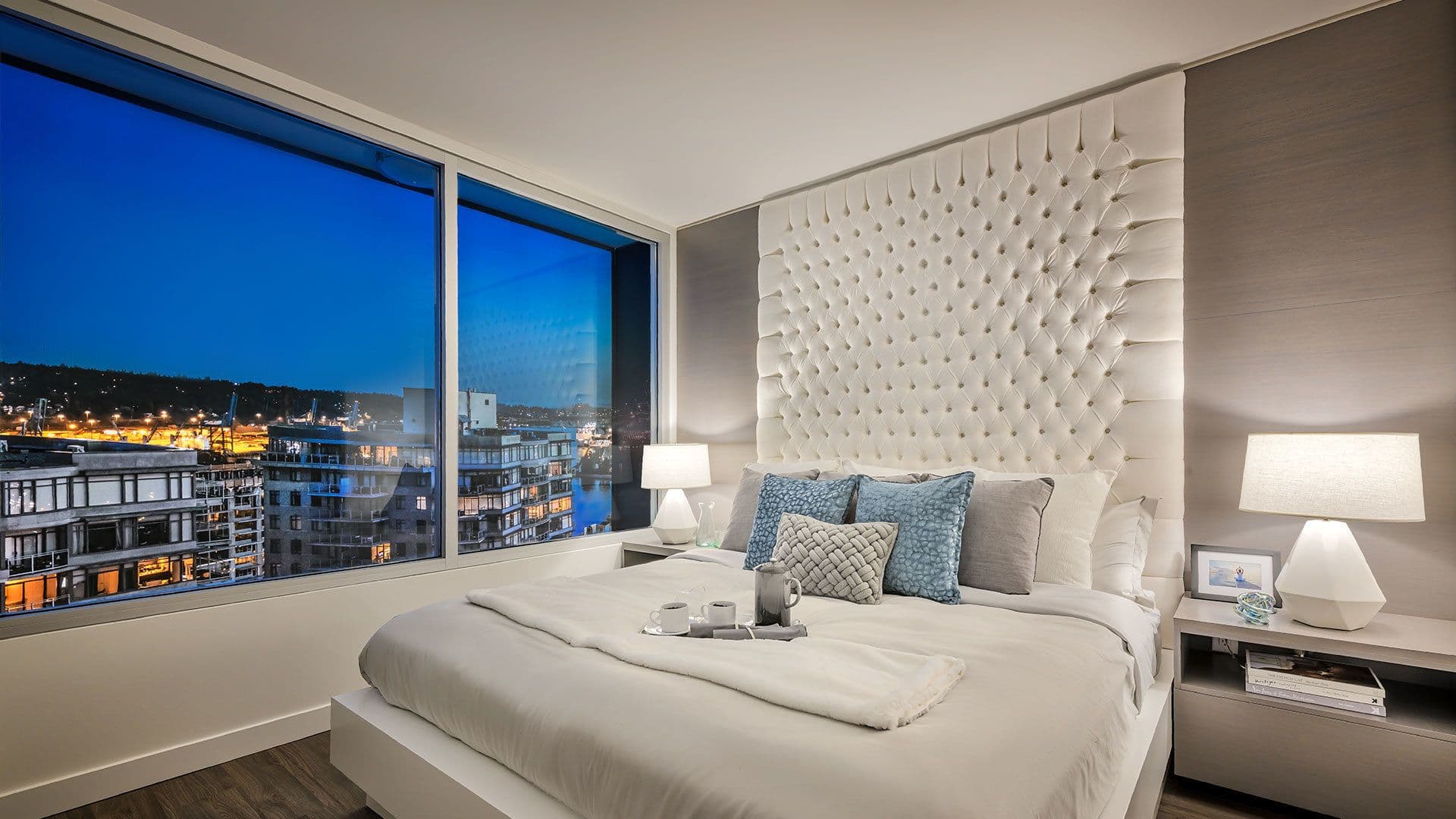 Ovation – A modern luxury bedroom featuring a large bed with a tall, white tufted headboard, flanked by matching nightstands with illuminated table lamps. Floor-to-ceiling corner windows showcase a dramatic dusk view of illuminated city buildings and glowing urban lights.