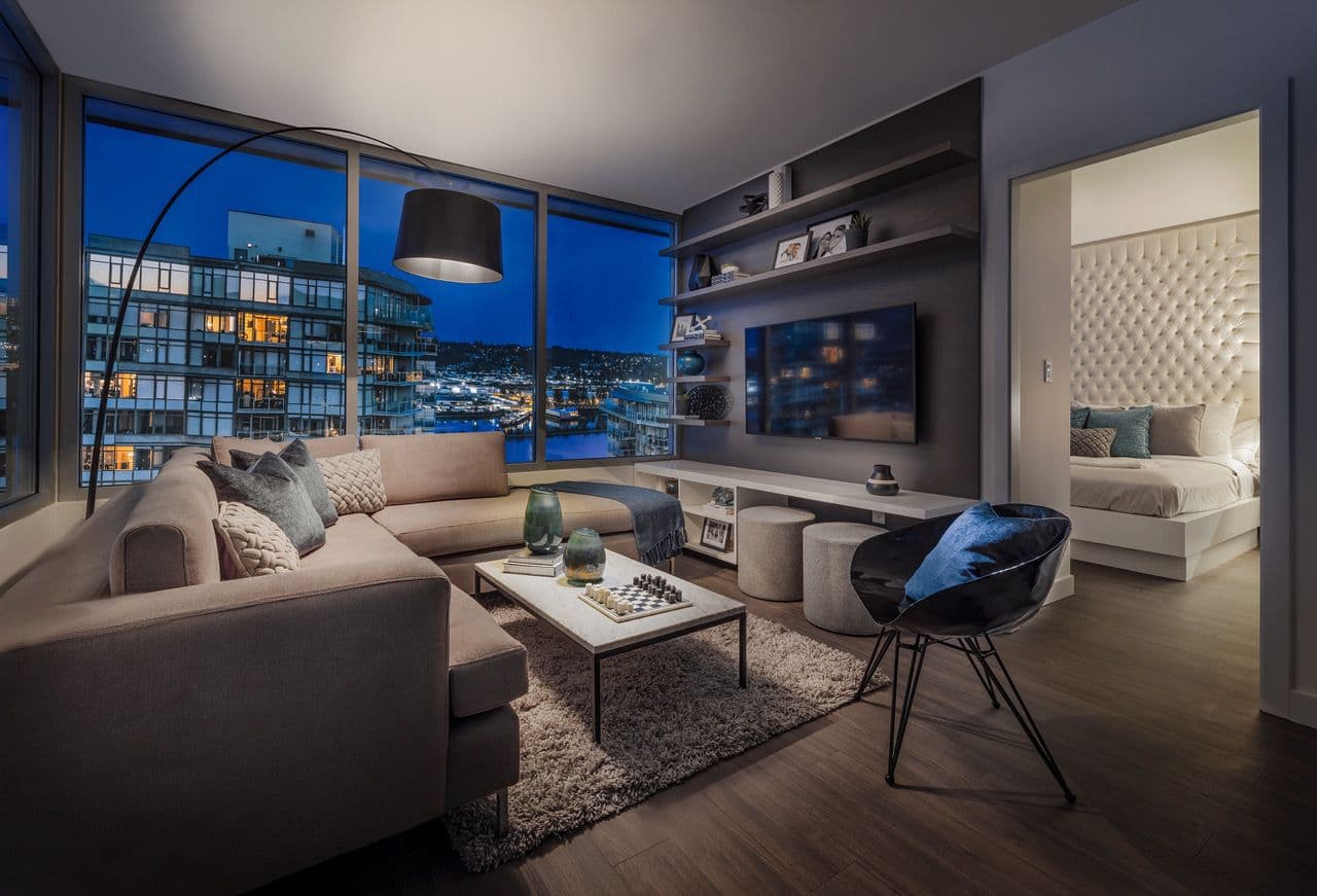 Ovation – A cozy, modern apartment living room features a beige sectional sofa, a dark accent wall with floating shelves around a mounted TV, and an open doorway leading to a bedroom with a tall tufted headboard. Large floor-to-ceiling windows showcase a evening city skyline view illuminated with city lights over a body of water.
