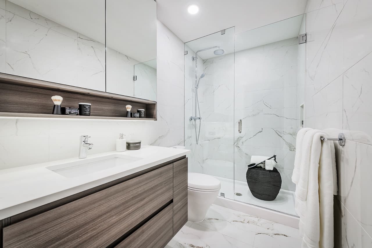 Ovation – A modern bathroom features floor-to-ceiling white marble tiles, a floating wood-finish vanity with a white countertop, and a sleek mirror cabinet above an open shelf. A glass-enclosed walk-in shower includes chrome fixtures and sits beside a white toilet under bright, clean recessed lighting.