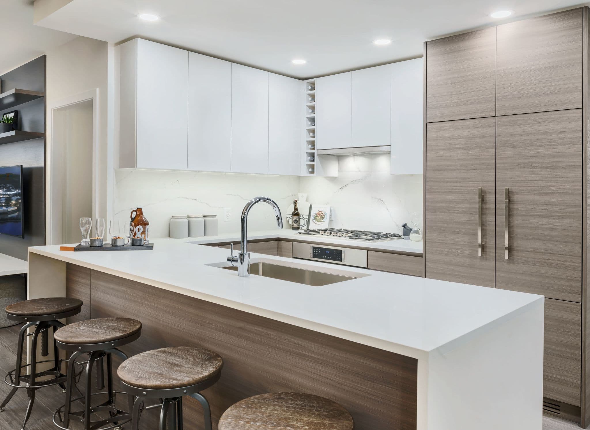Ovation – A modern kitchen featuring flat-panel white upper cabinets, light wood grain lower cabinets and full-height pantry panels, and crisp white quartz countertops with a marble-esque backsplash. An island with an undermount stainless steel sink and goose-neck faucet is lined with industrial-style barstools.