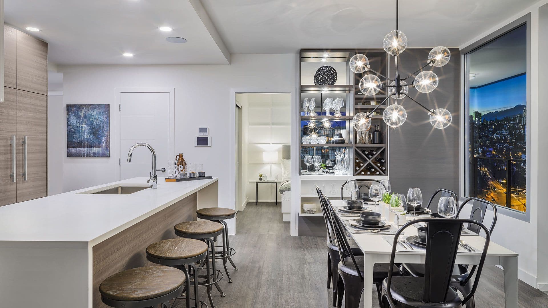 Ovation – A modern open-concept kitchen and dining area features a large island with barstools, wood-look cabinetry, and a set dining table under a contemporary globe chandelier. Large floor-to-ceiling windows reveal a glowing city skyline at dusk, while a custom lit bar niche adds elegance to the space.