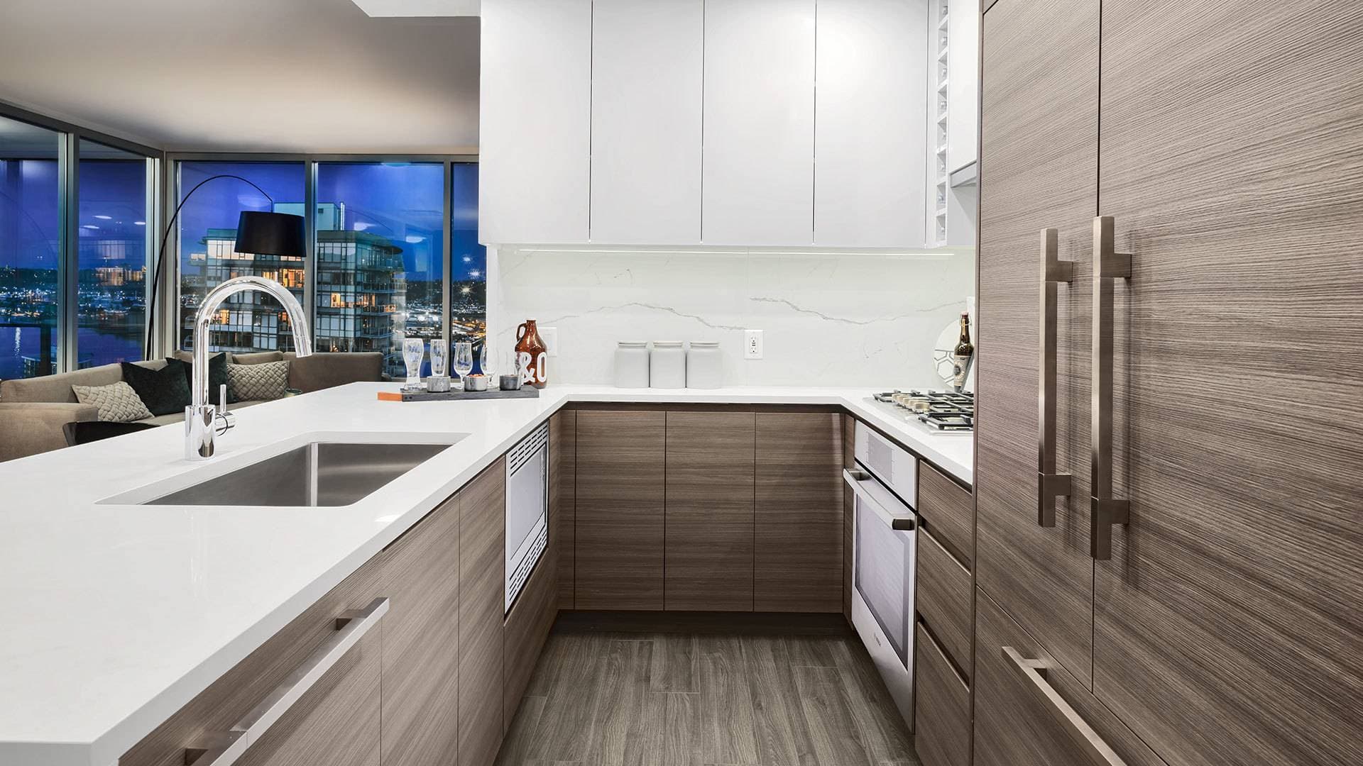 Ovation – A modern open-concept kitchen featuring flat-panel woodgrain lower cabinets, white upper cabinetry, and solid white quartz countertops with an undermount sink. Floor-to-ceiling windows in the adjacent living room frame a twilight view of city lights and surrounding towers.