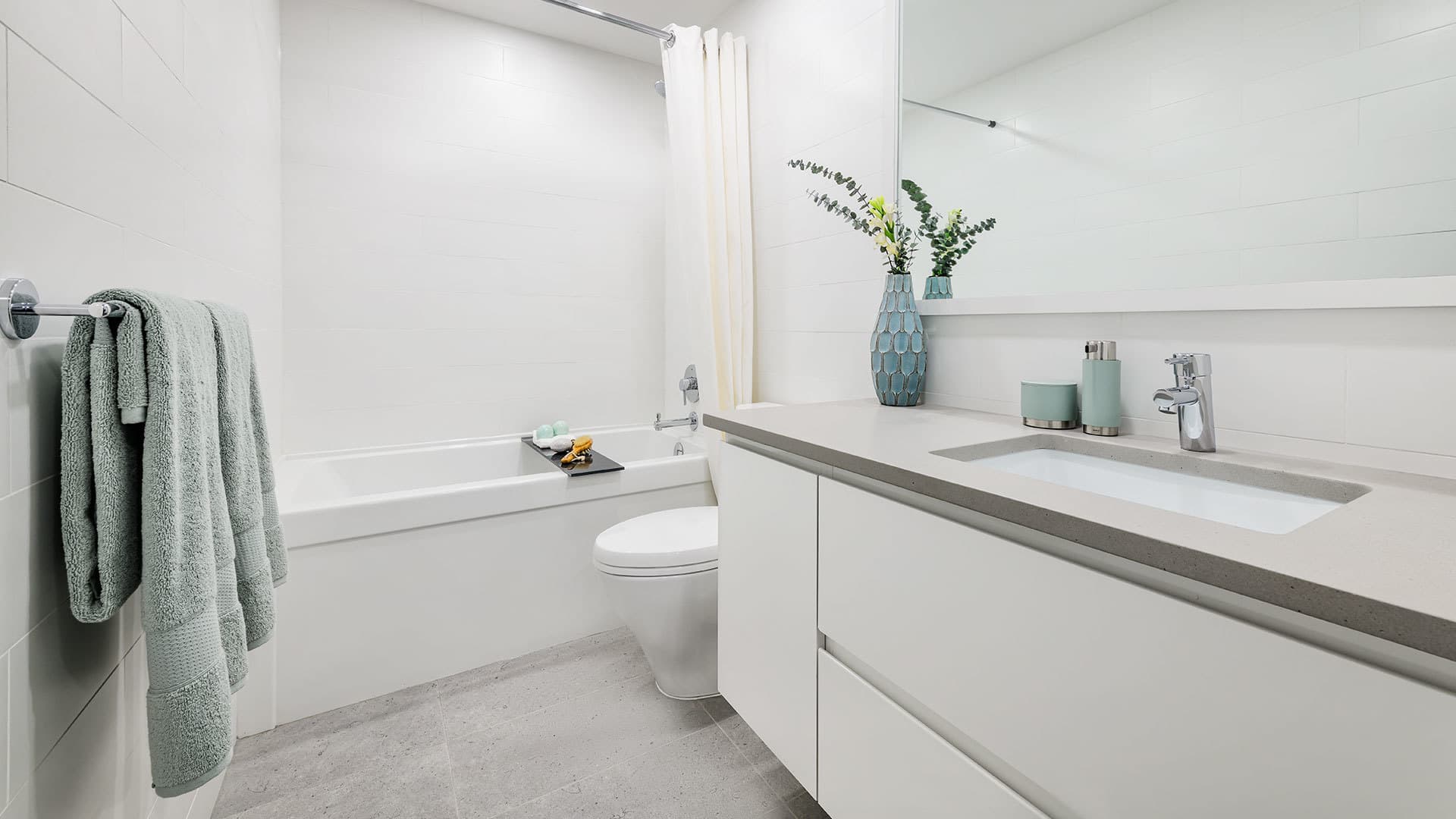 Ovation – A clean, modern apartment bathroom with bright white tiled walls and light grey floor tiles. The space features a floating white vanity with a dark quartz countertop, a large framed mirror, a modern toilet, and a white soaking tub with a shower curtain.