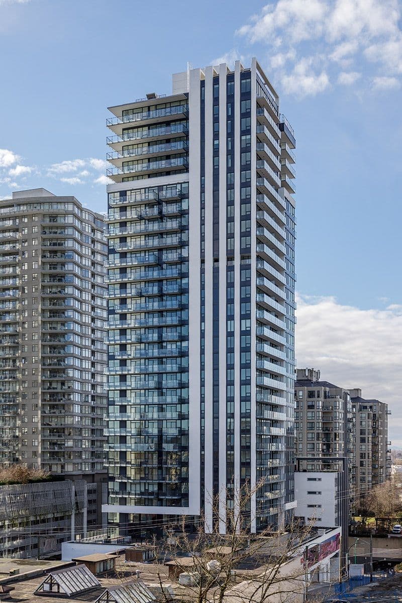 Ovation – A modern high-rise residential tower featuring prominent white vertical columns, dark-framed glass balconies, and extensive window walls stands against a bright blue sky with light clouds. Surrounding the main skyscraper are other contemporary condominium buildings and urban rooftops under crisp daytime sunlight.
