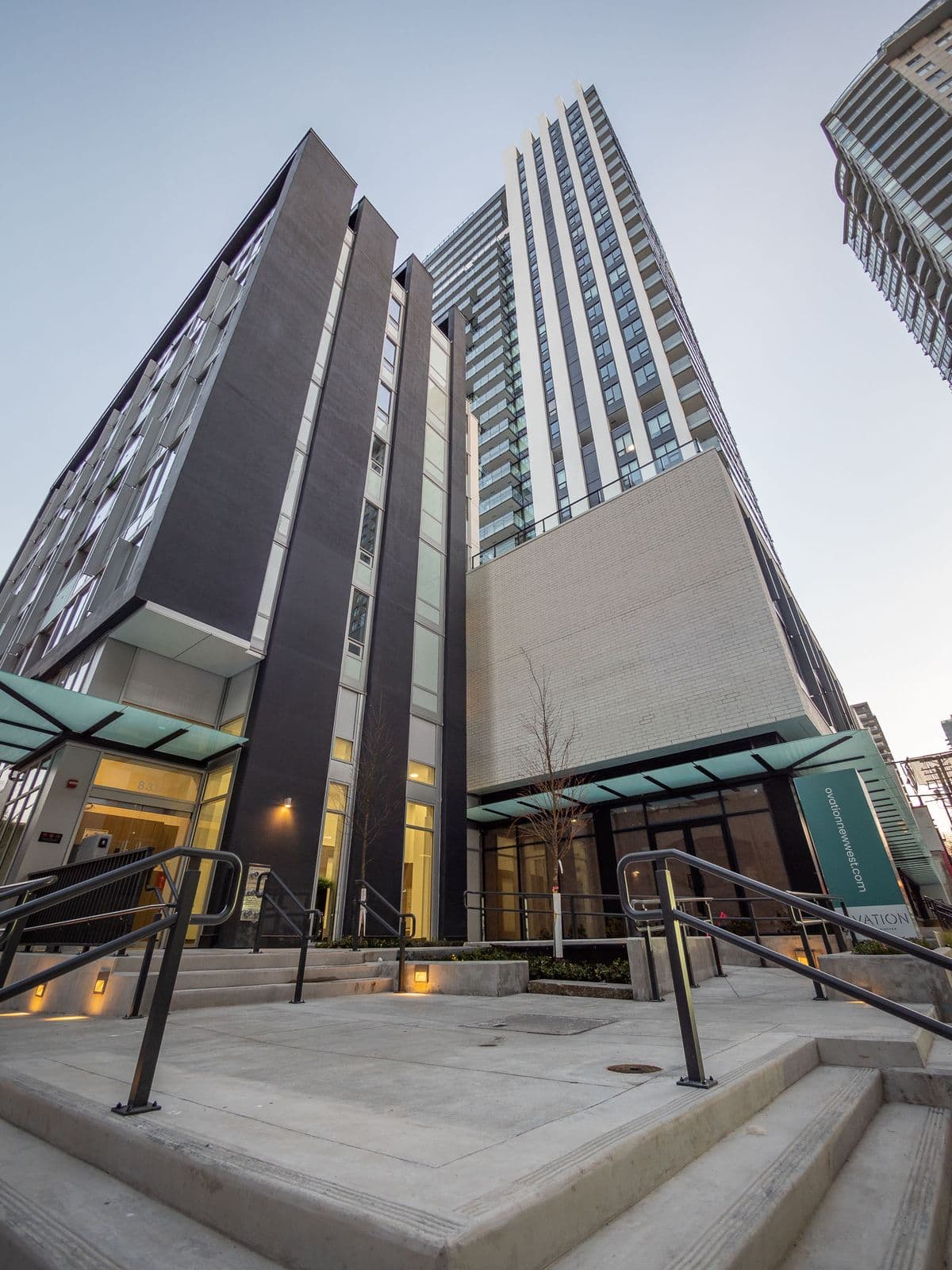 Ovation – Wide-angle upward view of the building's entrance area showing two connected towers with dark and light facades and glass canopy entries.