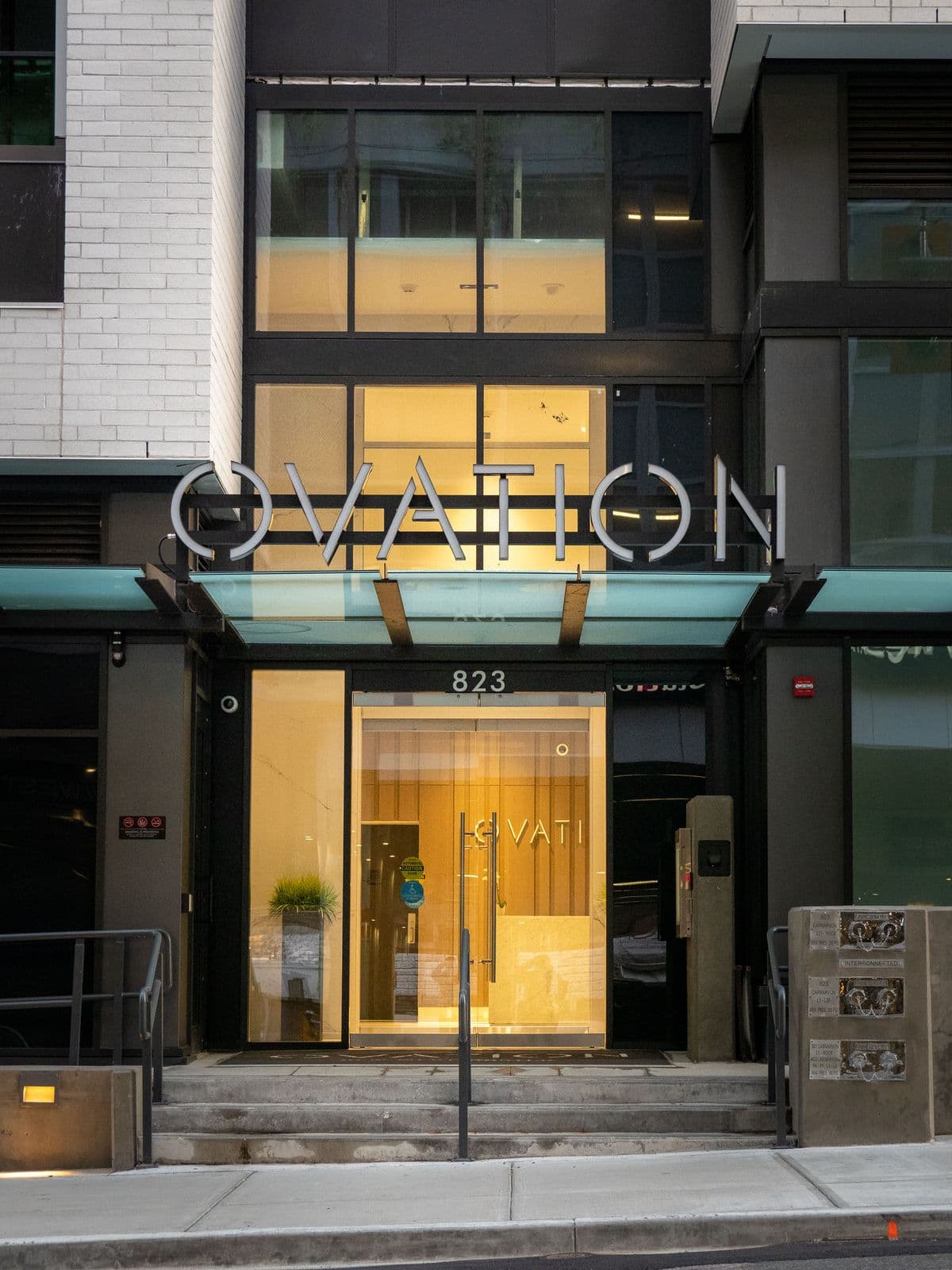 Ovation – Close-up of the building's main entrance at dusk with an illuminated lobby, glass doors, and an 'OVATION' sign above the canopy.
