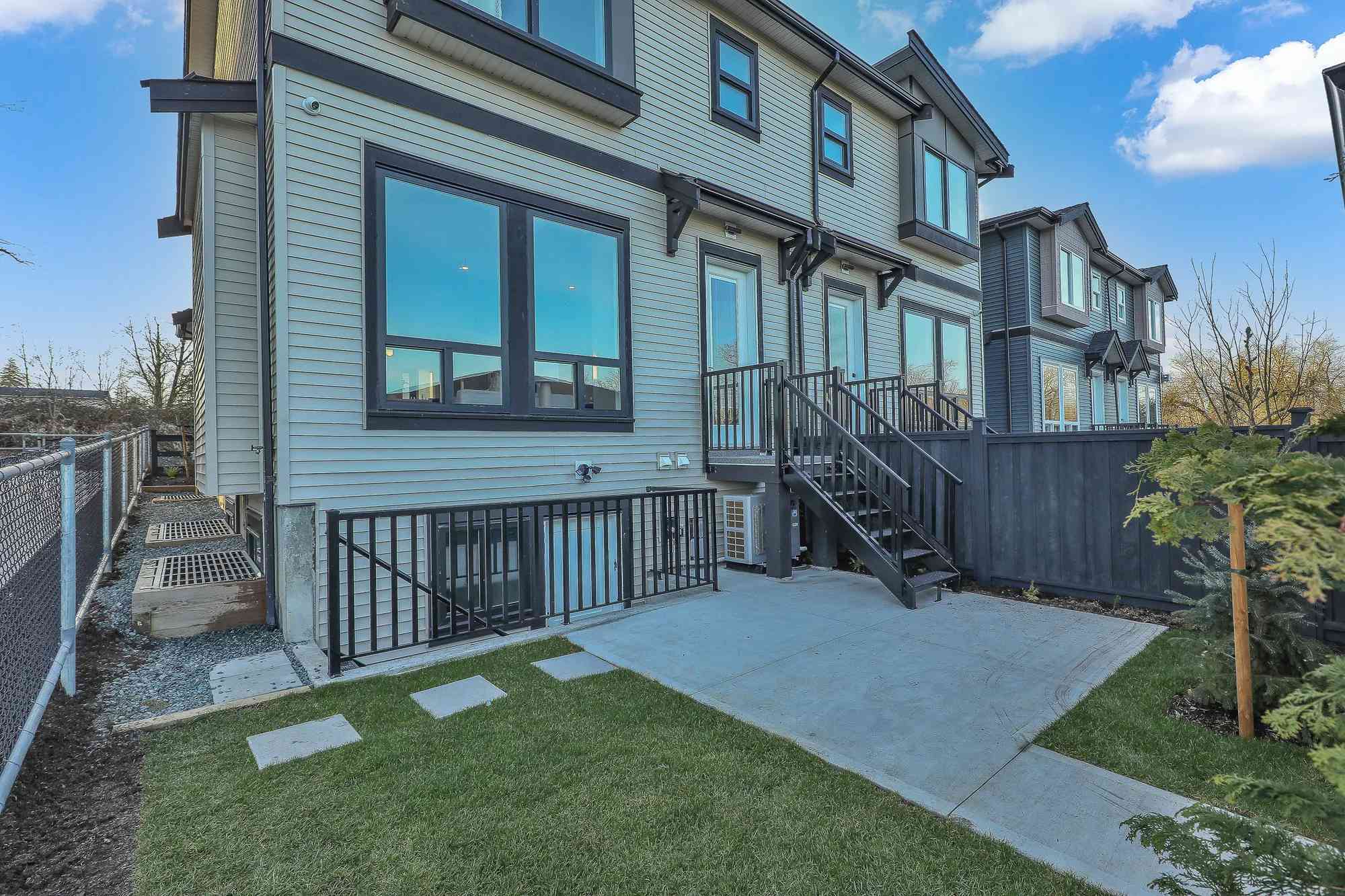 Latimer Landing – The rear exterior of a multi-story townhouse clad in light horizontal siding, featuring dark-trimmed windows and an outdoor metal staircase leading to a backyard patio. A small patch of green lawn, dark fencing, and clear blue skies with light clouds complete the residential scene.