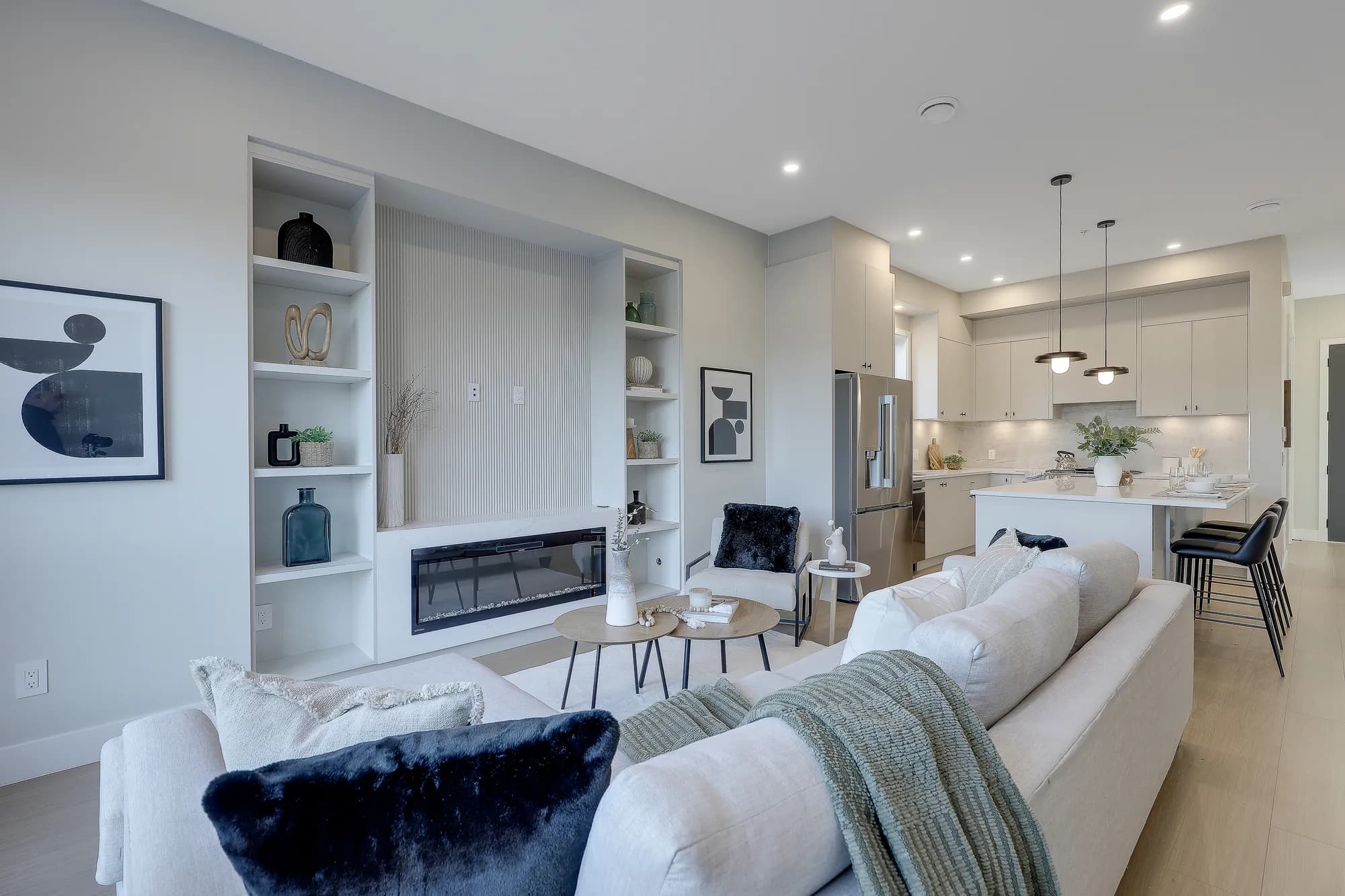Latimer Landing – A modern open-concept living room and kitchen featuring a built-in entertainment unit with vertical fluting, integrated shelving, and a horizontal linear fireplace. The space is styled with neutral-toned sectional seating, accent pillows, round nesting coffee tables, and view into the kitchen equipped with flat-panel cabinetry and stainless steel appliances.