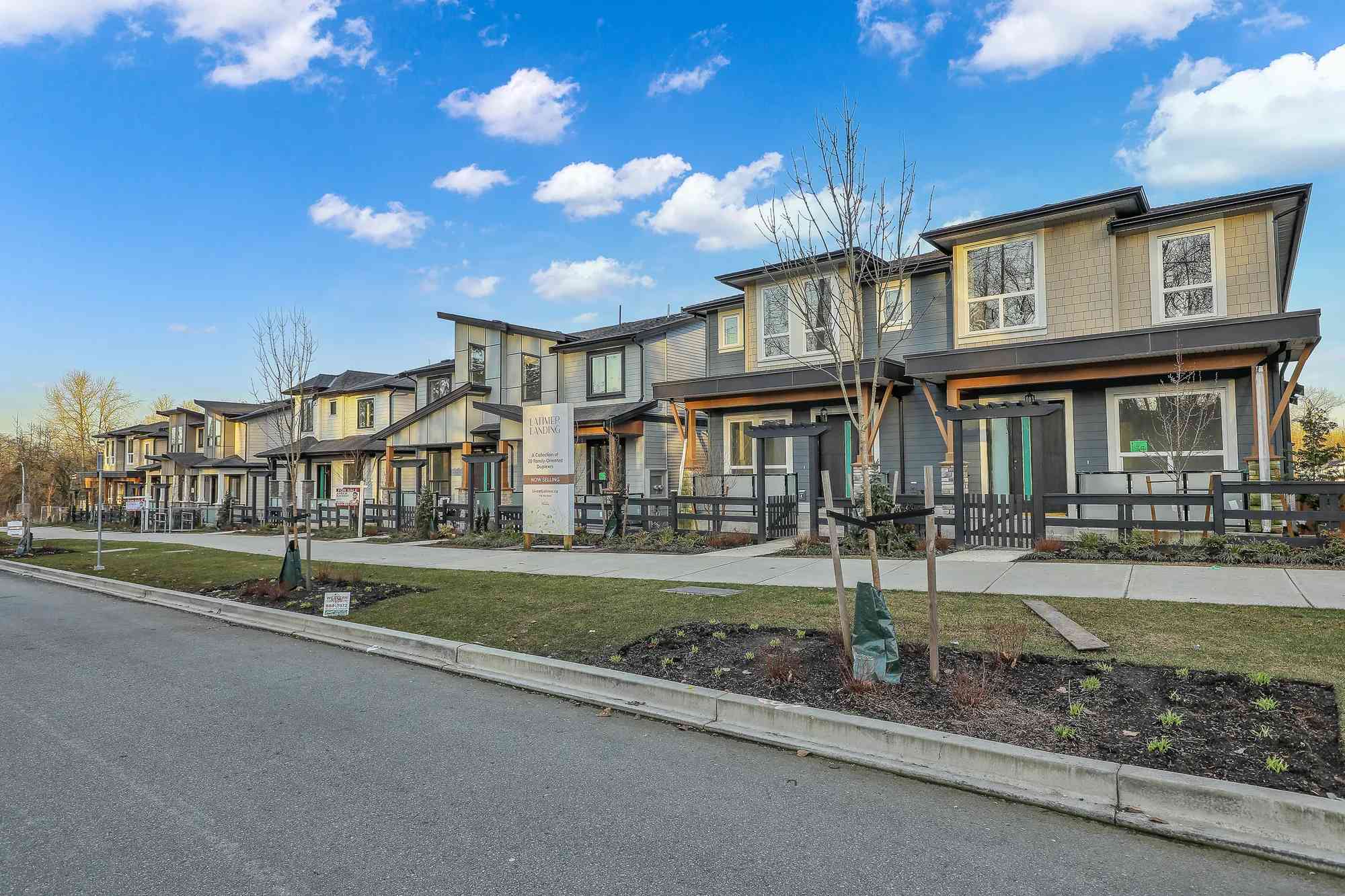 Latimer Landing – A row of newly constructed, modern two-story townhomes lines a quiet residential street, featuring a mix of grey siding, warm wood accents, and low black picket fences. Young saplings in support stakes border the paved sidewalk and grass boulevards beneath a clear blue sky dotted with soft white clouds.