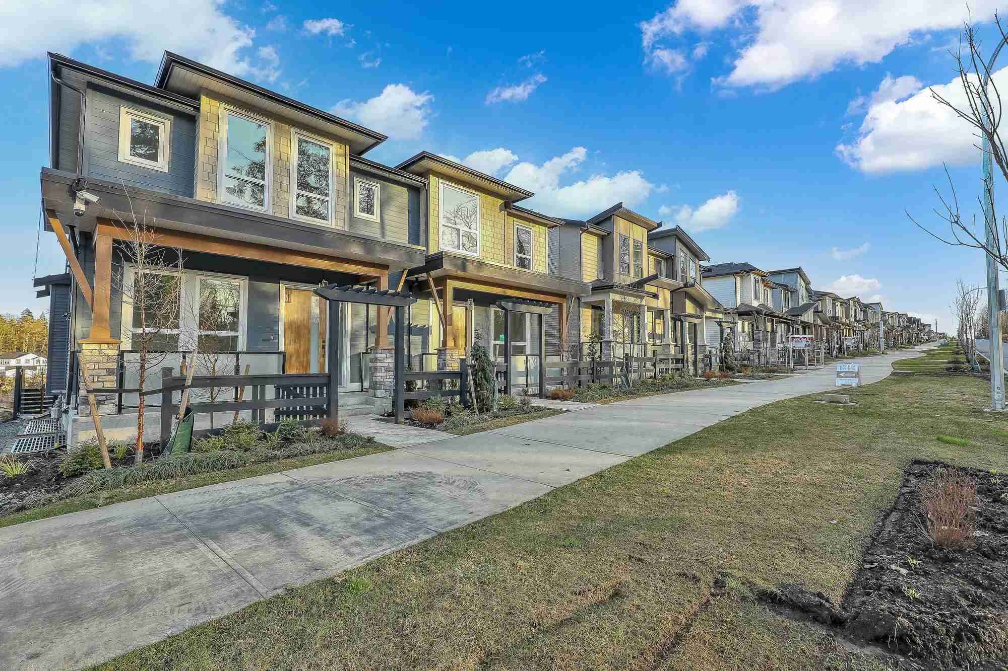 Latimer Landing – A wide daytime shot of a long row of modern multi-level townhomes featuring stone accents, warm wood posts, and mixed siding in grey and yellow tones. A clean concrete sidewalk and landscaped front yards with low fencing run parallel to the homes beneath a bright blue, partly cloudy sky.