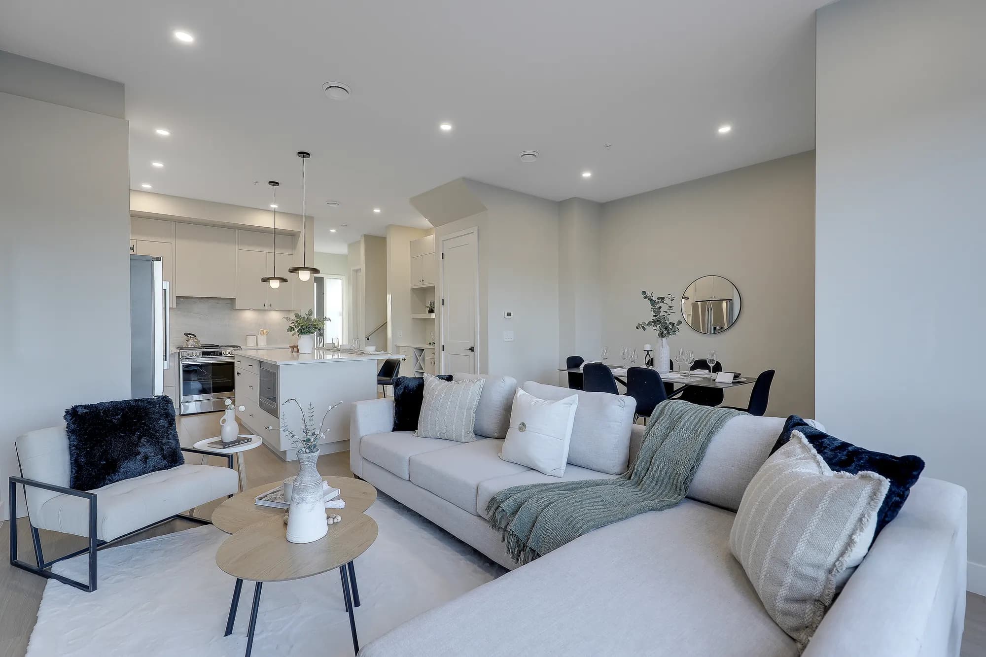 Latimer Landing – An open-concept living area featuring a large, light grey sectional sofa with decorative pillows, nested wood coffee tables, and an accent armchair on a soft rug. In the background, the space flows seamlessly into a dining area and a modern kitchen equipped with an island, sleek off-white cabinetry, and stainless steel appliances.