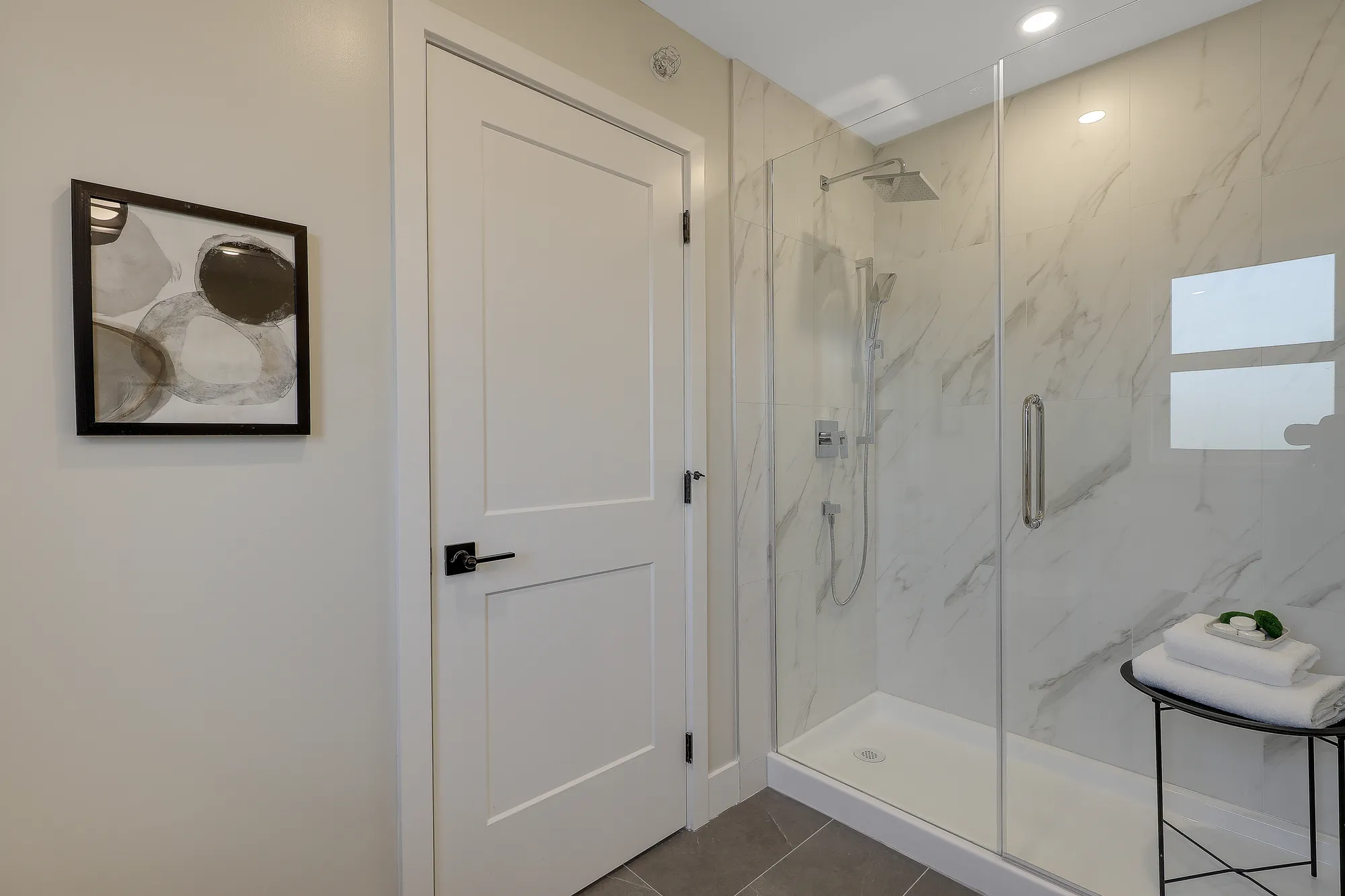 Latimer Landing – A bright modern bathroom featuring a white paneled door with black hardware beside a walk-in shower enclosed in seamless glass. The shower is lined with white marble-patterned tiles and equipped with chrome fixtures, complemented by dark floor tiling and a small side table holding folded towels.