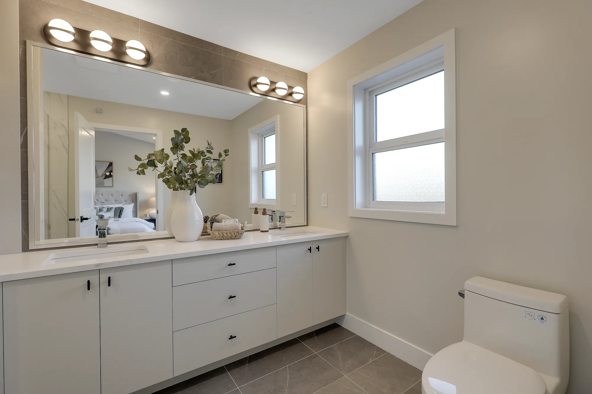 Latimer Landing – A bright modern bathroom featuring a long white double vanity with modern black hardware and a large wall-spanning mirror illuminated by sconce lighting. A frosted privacy window brings in natural light, while the mirror reflects an adjoining bedroom with a made bed.