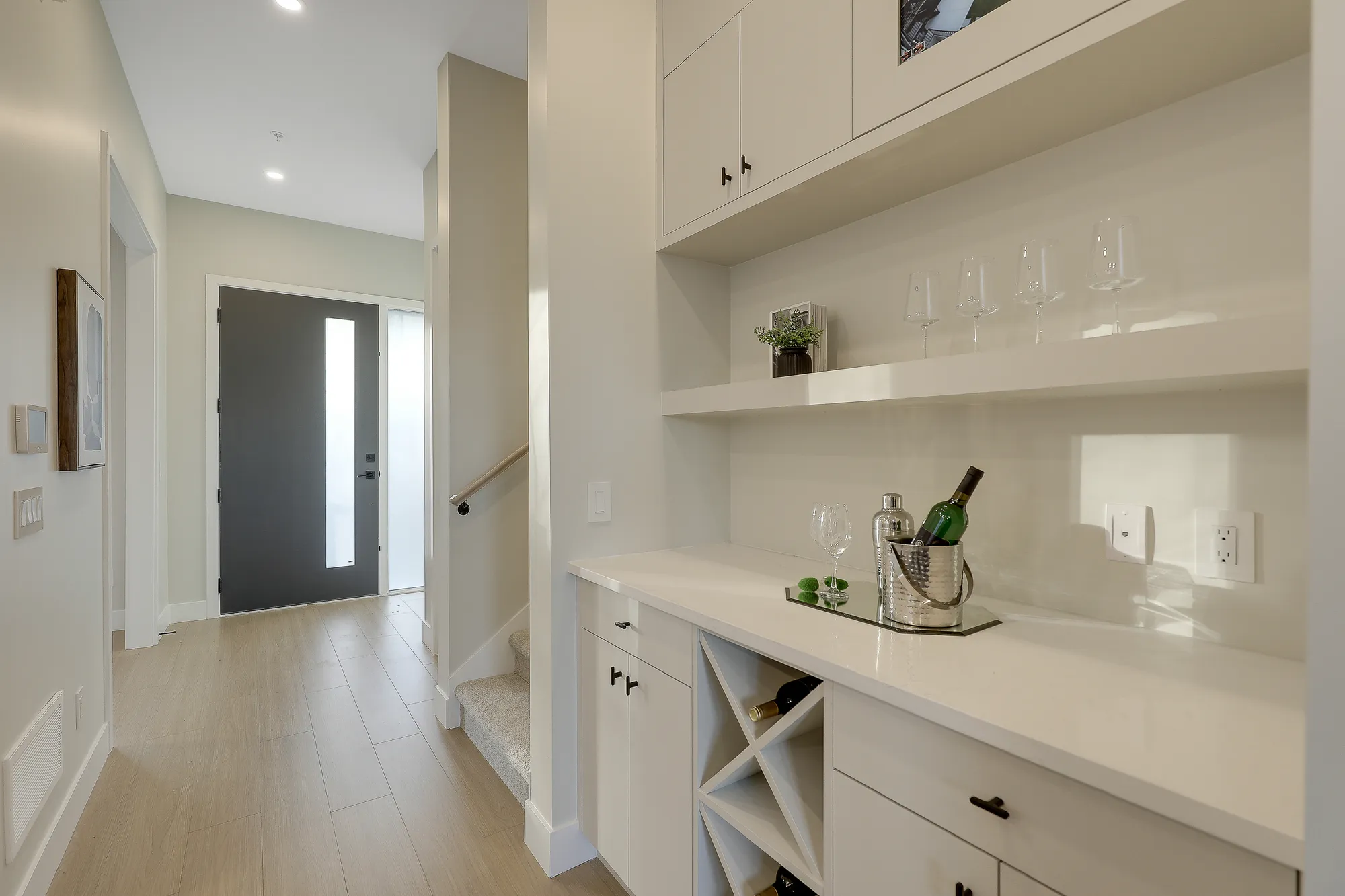 Latimer Landing – A bright home entryway hallway featuring light wood flooring, with a built-in wet bar and wine station on the right equipped with white cabinetry, open shelving, and a wine bottle chiller. In the background, the dark modern front door with frosted vertical glass sits alongside a small staircase leading to an upper level.