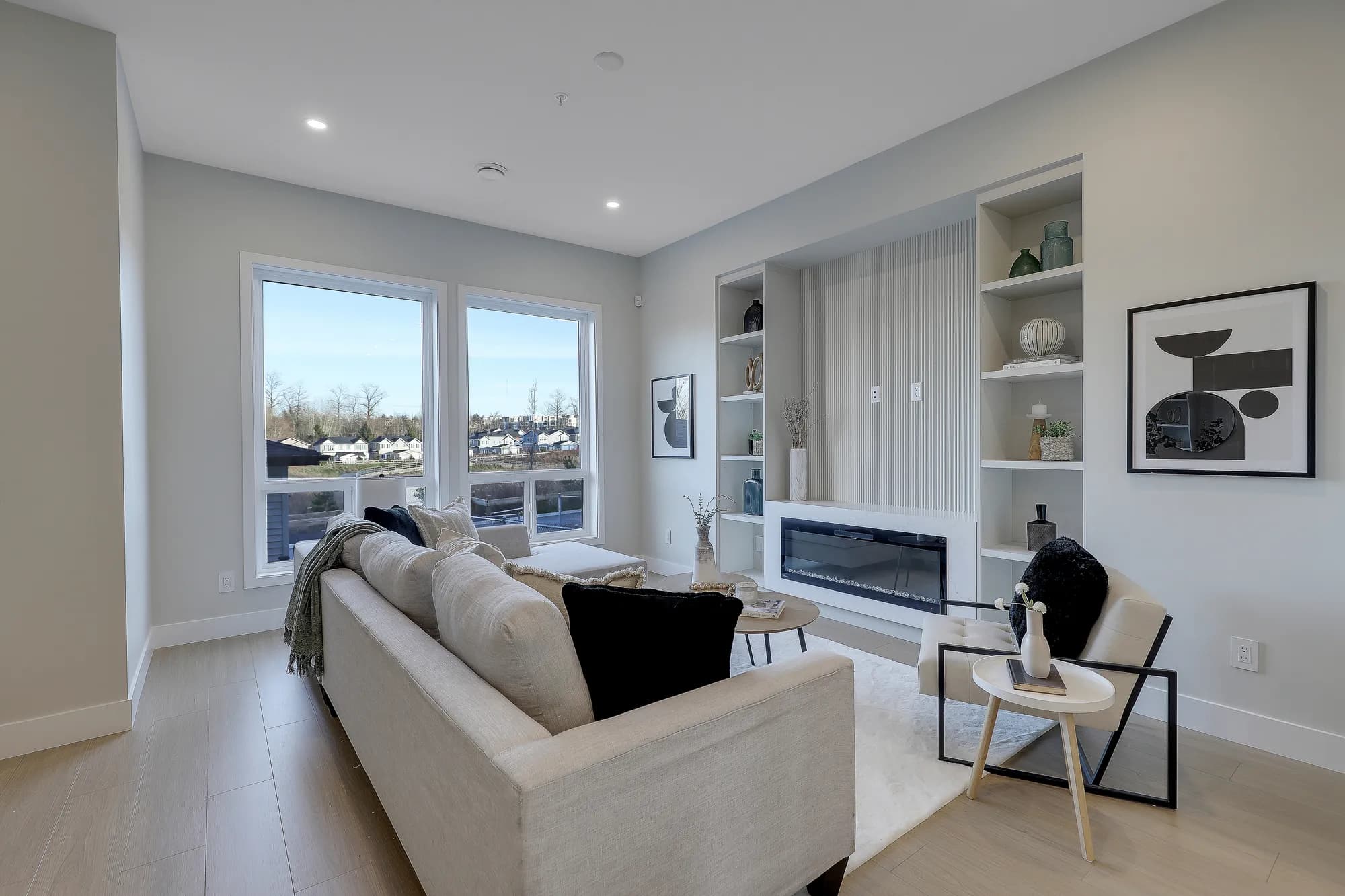 Latimer Landing – A modern living room features a built-in media wall with open display shelving, a ribbed panel backdrop, and an integrated linear electric fireplace. The space is furnished with light-toned seating, a plush white rug, and light hardwood floors, naturally illuminated by large windows offering daytime views of a neighboring residential community.