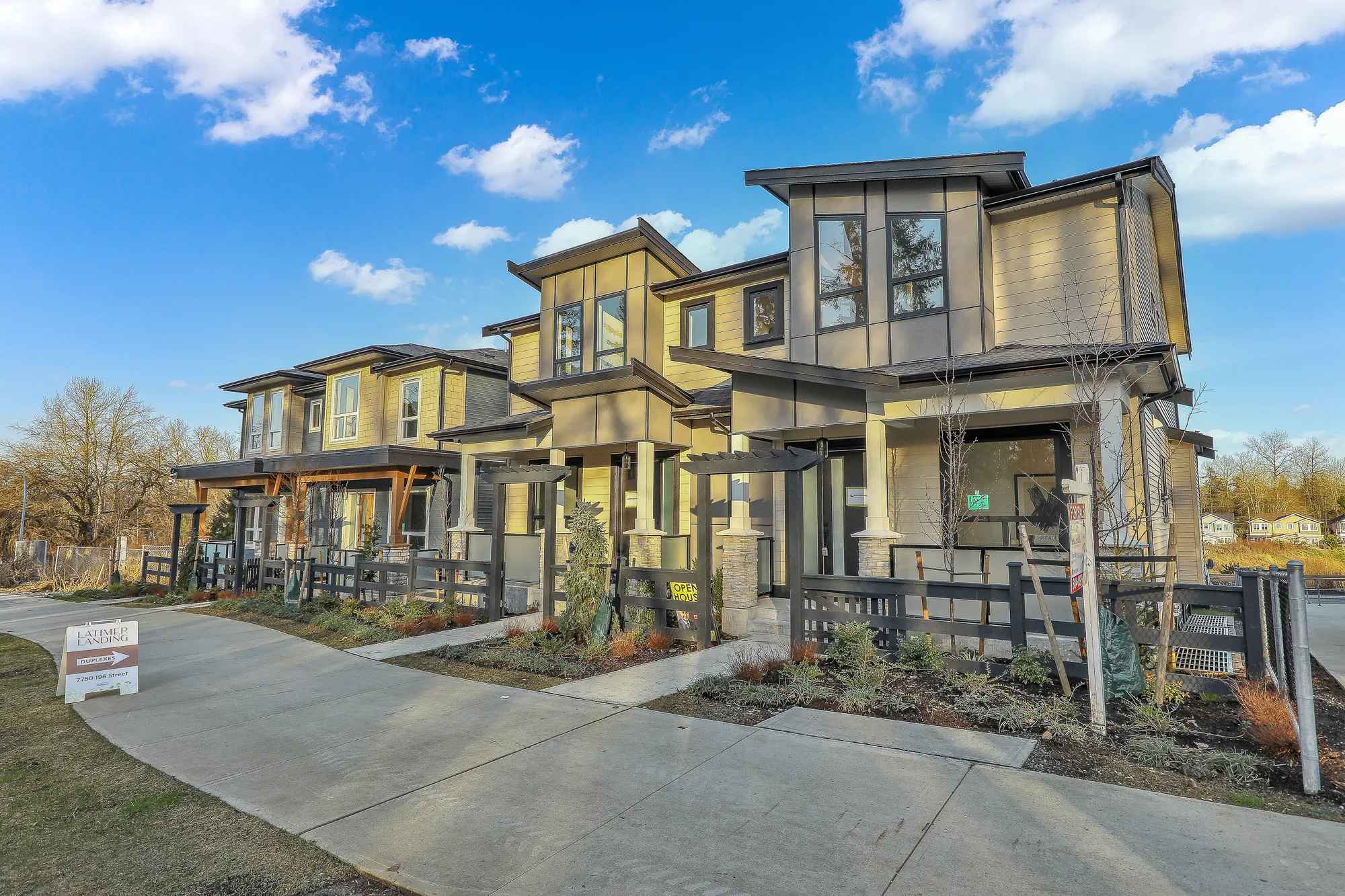 Latimer Landing – A ground-level view shows a row of newly constructed, modern two-story duplex homes featuring beige and grey siding, dark trim, and covered front porches with stone-accented pillars. Concrete pathways run alongside dark fenced front yards with fresh landscaping under a bright blue, cloud-dotted sky.