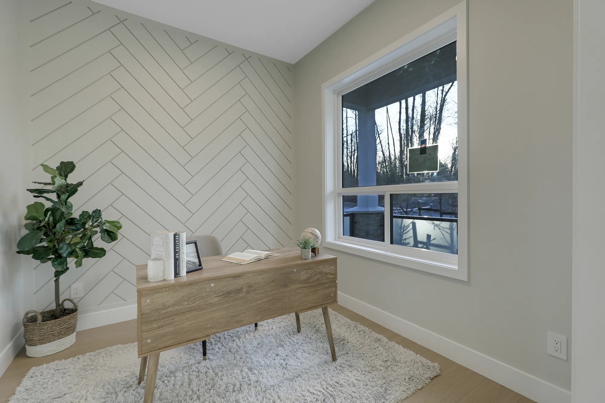 Latimer Landing – A bright home office room features a light wood mid-century modern desk situated on a plush shag rug. A patterned chevron accent wall, an indoor potted plant, and a large window looking out onto a wooded setting complete the quiet workspace.